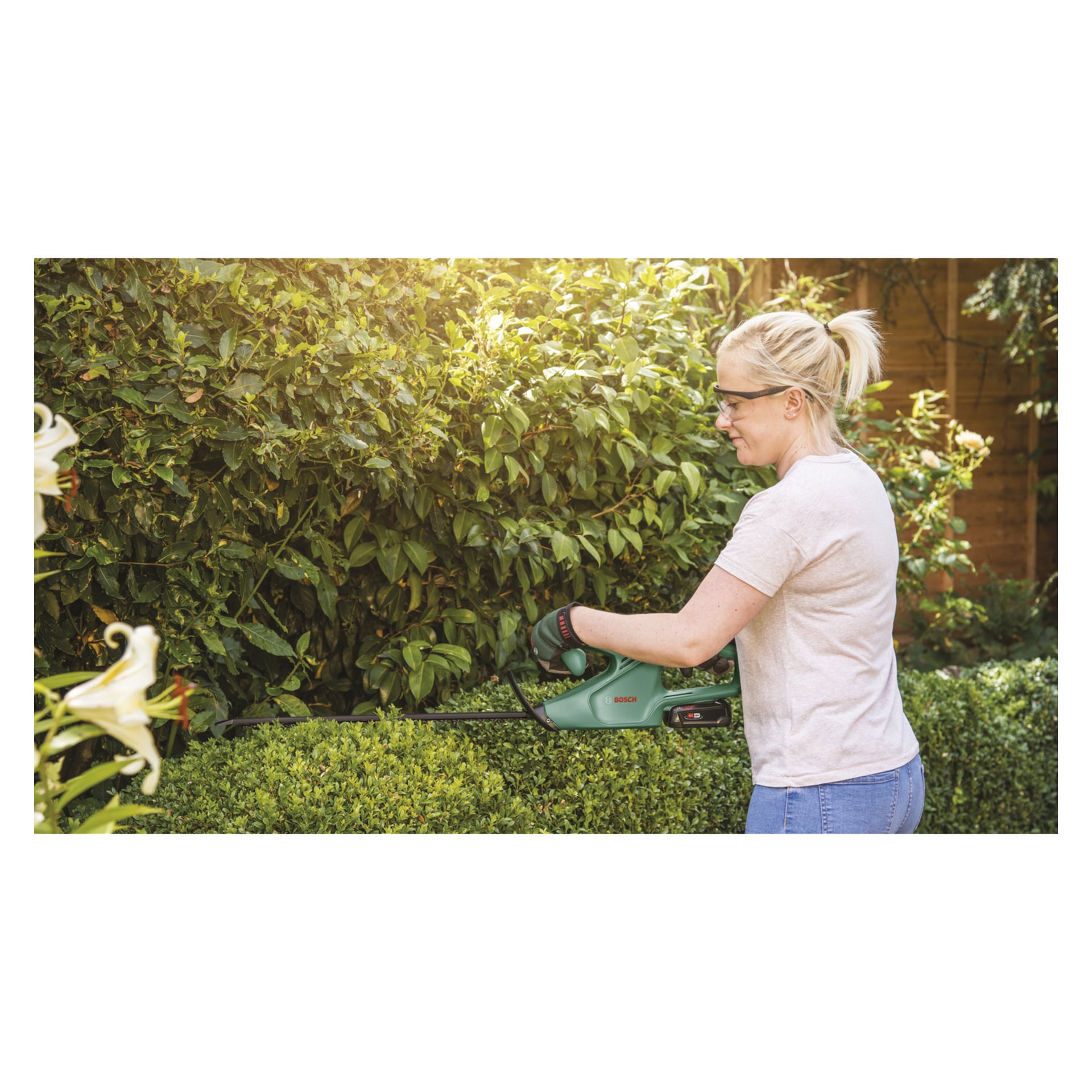 Någon med skyddsglasögon och handskar klipper en häck med en Bosch Power 4 All EasyHedgeCut 18-45 batteridriven trimmer, omgiven av gröna buskar och blommande blommor.