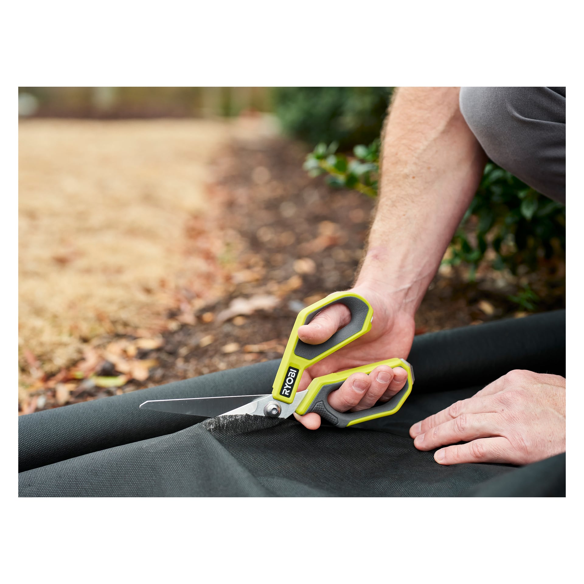 Någon klipper svart trädgårdsduk nära täckmaterial och jord med gröna och grå Ryobi Scissors RHCSS01. En hand säkrar tyget, medan den andra hanterar saxen. Det finns buskar och gräs synliga i bakgrunden.