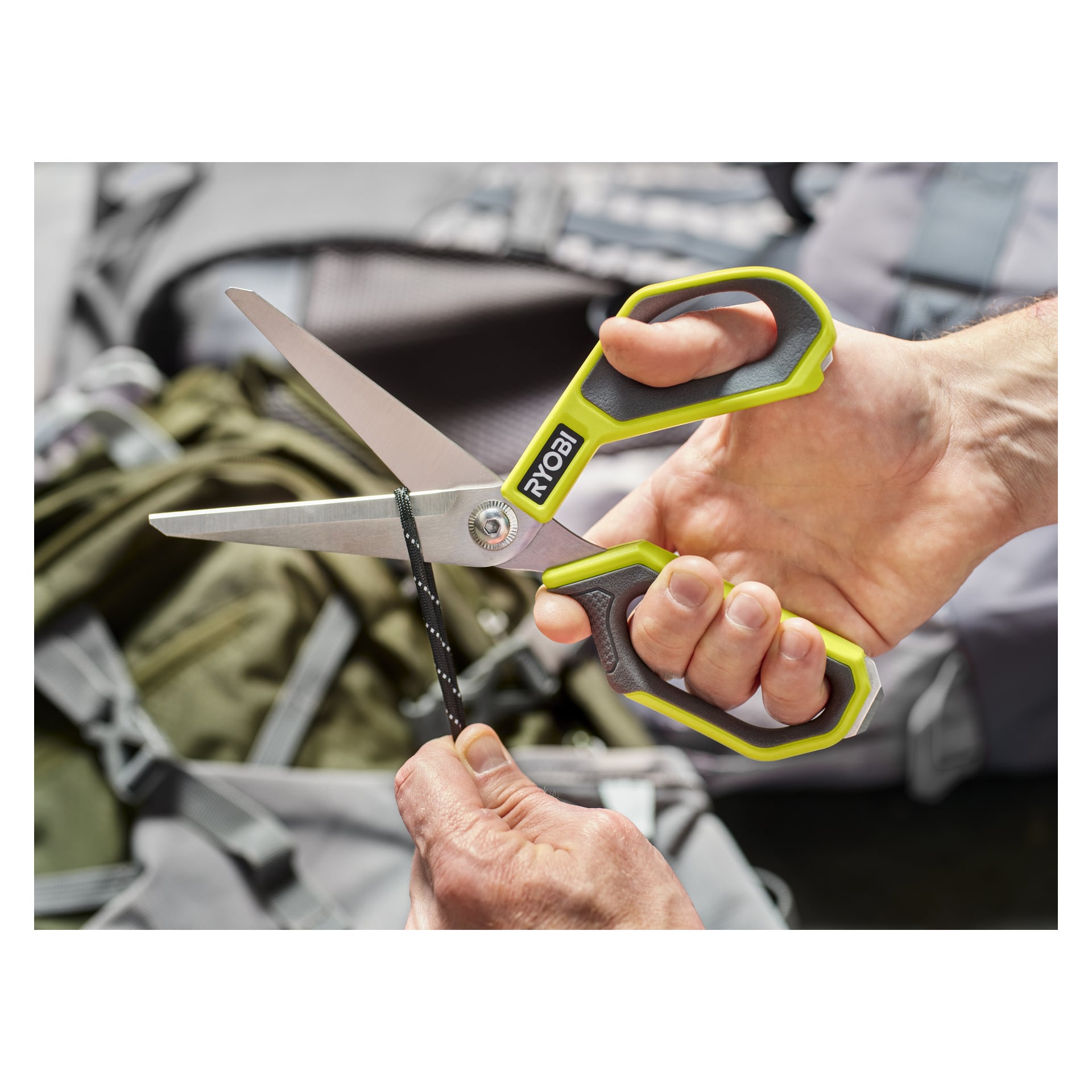 Någon klipper en bit svart rep med grönskaftade Ryobi Scissors RHCSS01, och ryggsäckar och väskor syns suddiga i bakgrunden.
