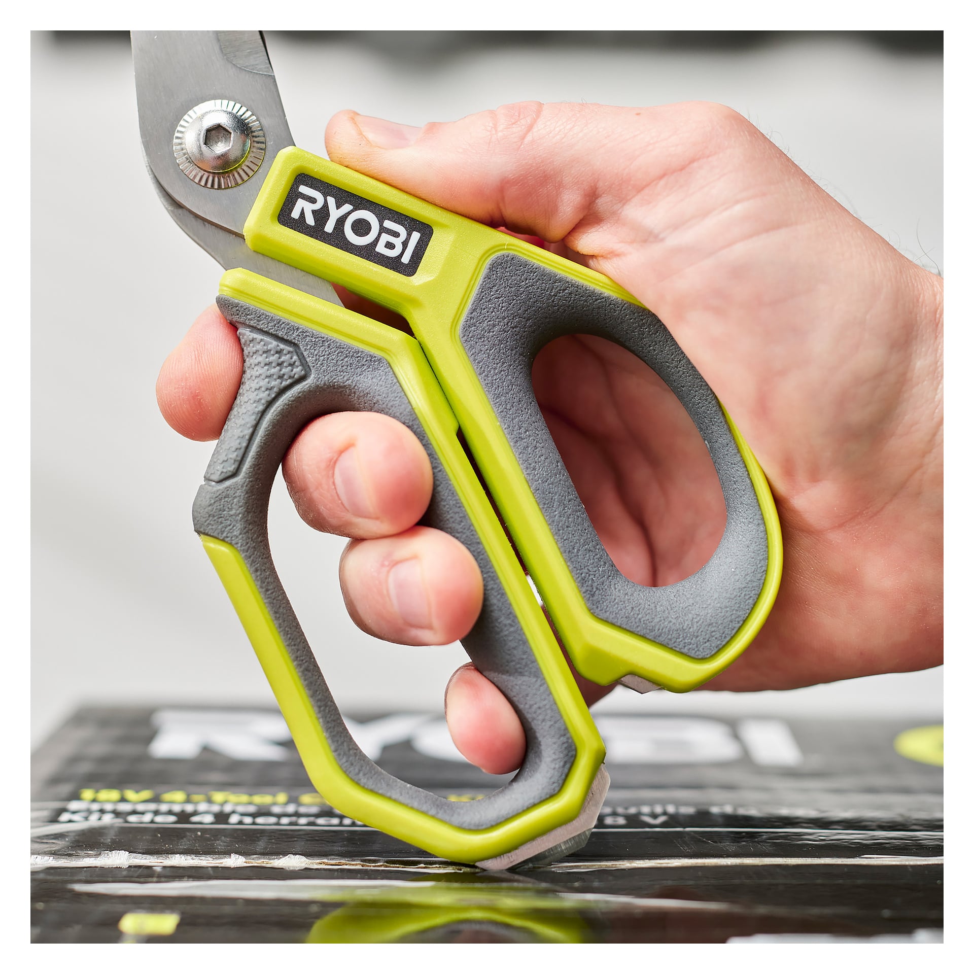 En hand greppar Ryobi Scissors RHCSS01, som har ett grönt och grått handtag med Ryobi-loggan. Bakgrunden är suddig och innehåller en del av Ryobis förpackning.