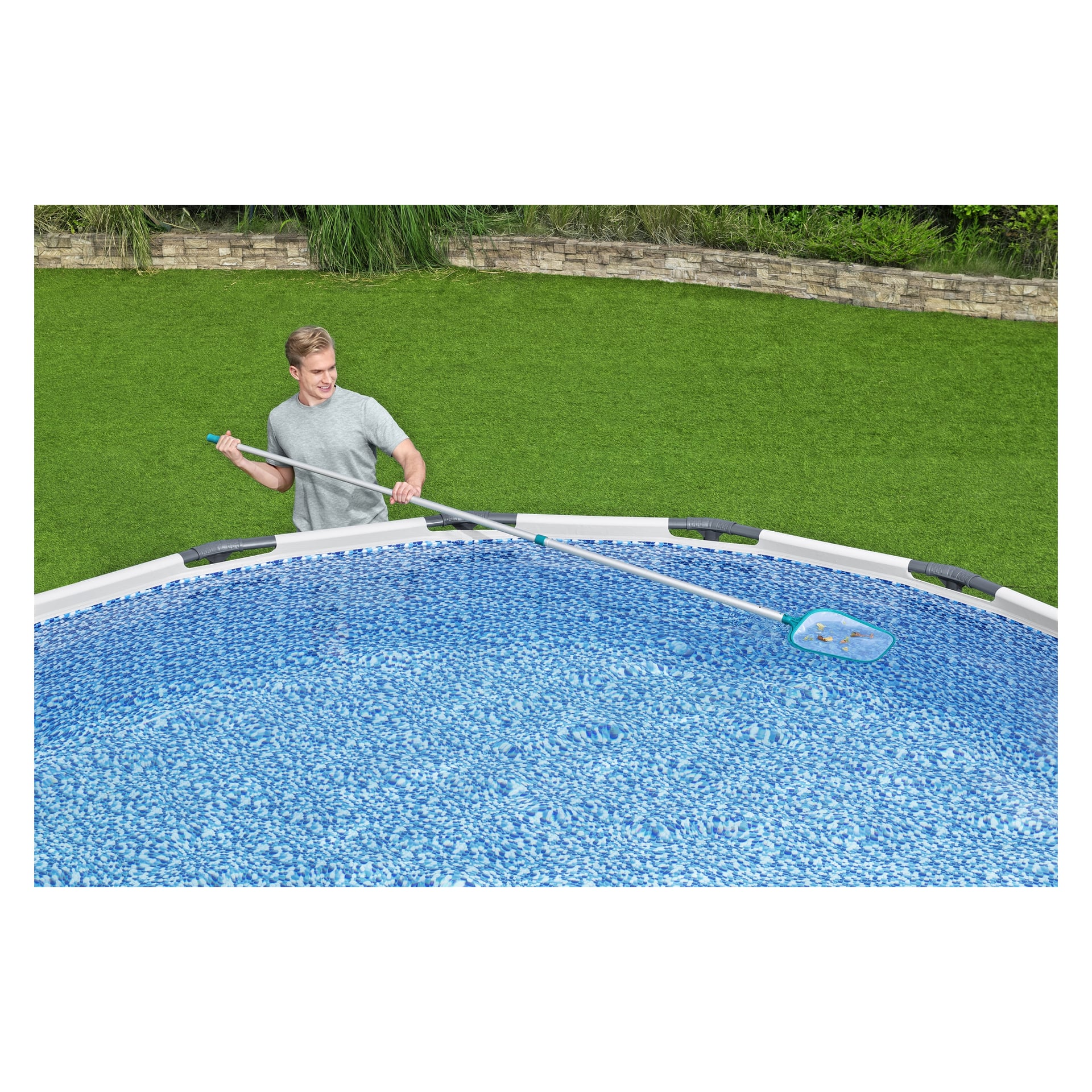 Någon använder Bestway AquaClean 3-delade rengöringsset med ett långt handtag för att rengöra skräp från en ovanjordisk pool. Området runt poolen har grönt gräs och en stenmur kan ses i bakgrunden.