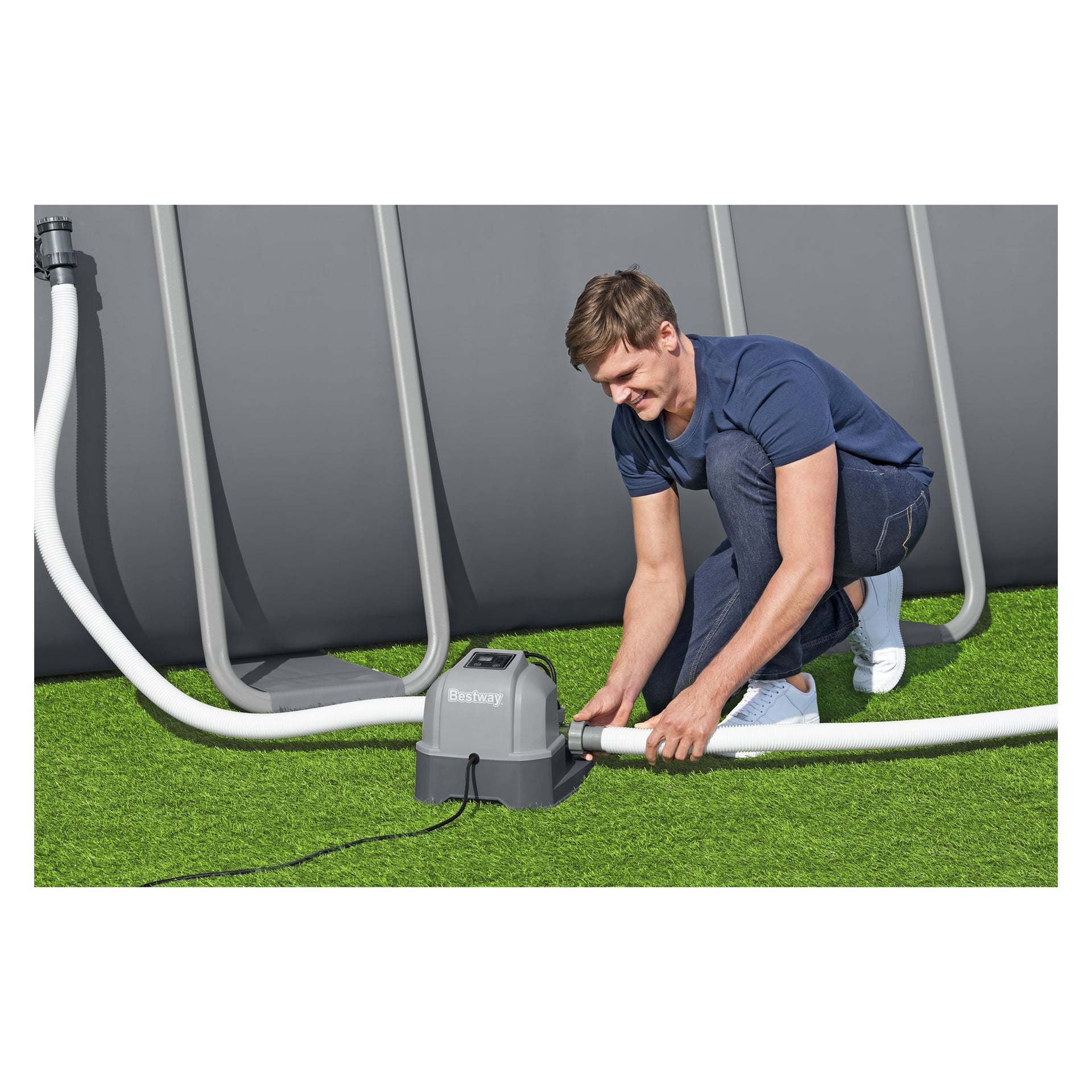 En person i blå skjorta och jeans knäböjer på konstgräs och kopplar en slang till det grå Bestway Salt Chlorinator-systemet bredvid en ovanjordisk pool.