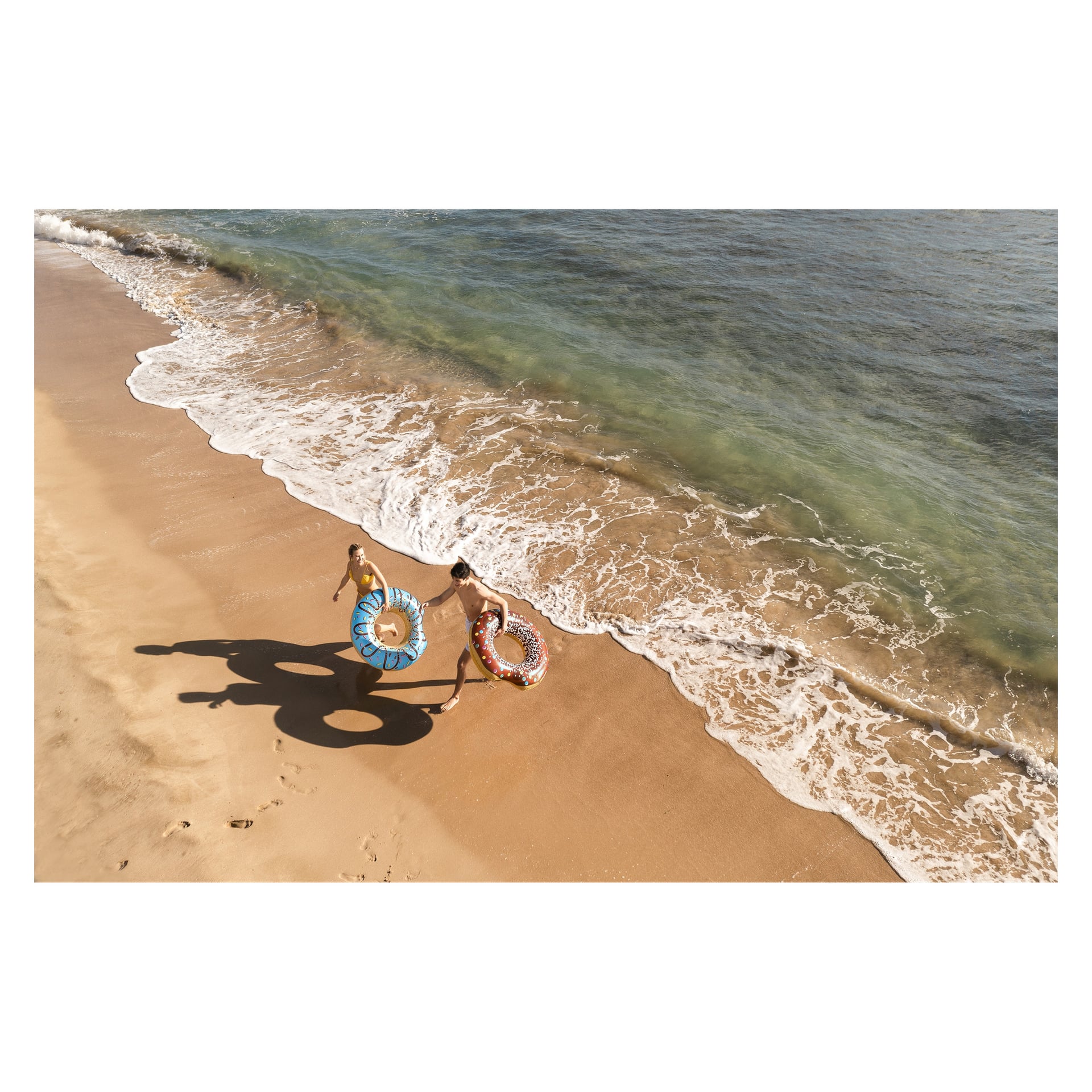 Två personer går sida vid sida längs en sandig strandlinje, var och en håller en en färgstark Bestway Swim-ring. Deras skuggor följer efter dem på den våta sanden medan mjuka havsvågor rullar in.