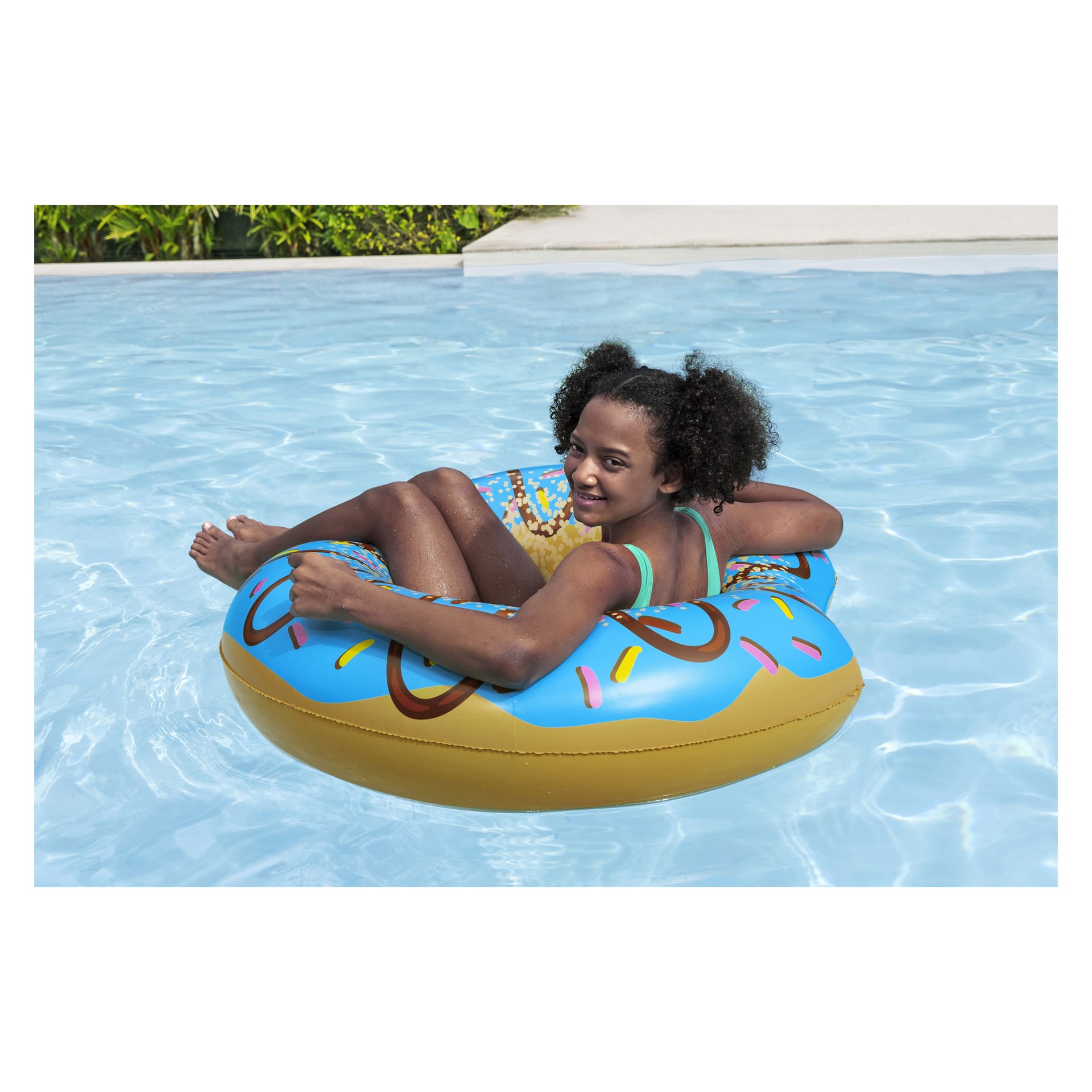 En lockhårig flicka sitter i en Bestway Swim-ring och flyter i en kristallklar blå pool och tittar tillbaka med ett leende. Bakgrunden har frodig grönska och en del av pooldäcket.