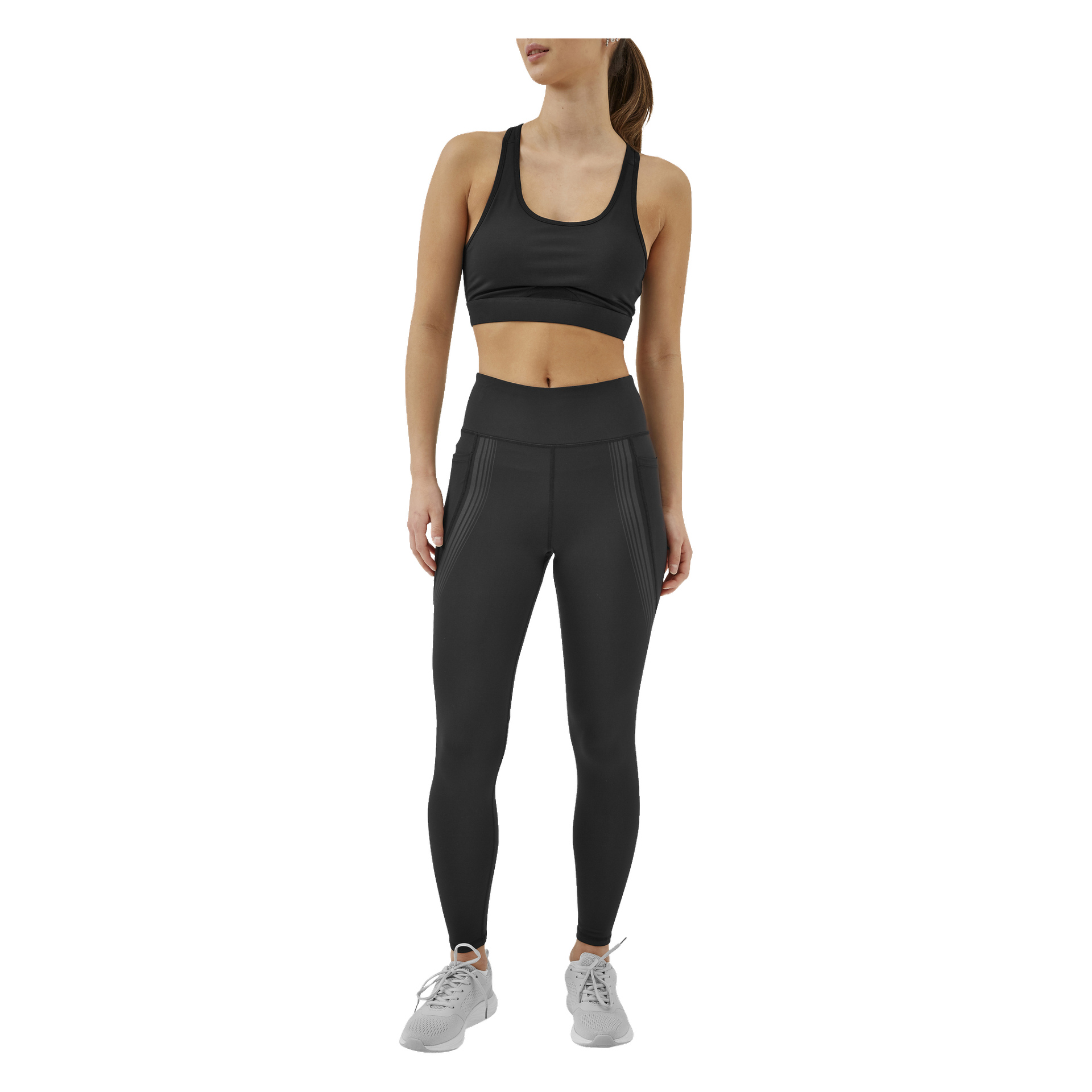 En kvinna modellerar Kayoba Tights damkläder i Boston, klädd i högmidjade svarta leggings med diskreta ränder och en matchande svart sport-bh. Hon matchar sin outfit med ljusgrå träningsskor och har håret uppsatt, lite åt sidan.