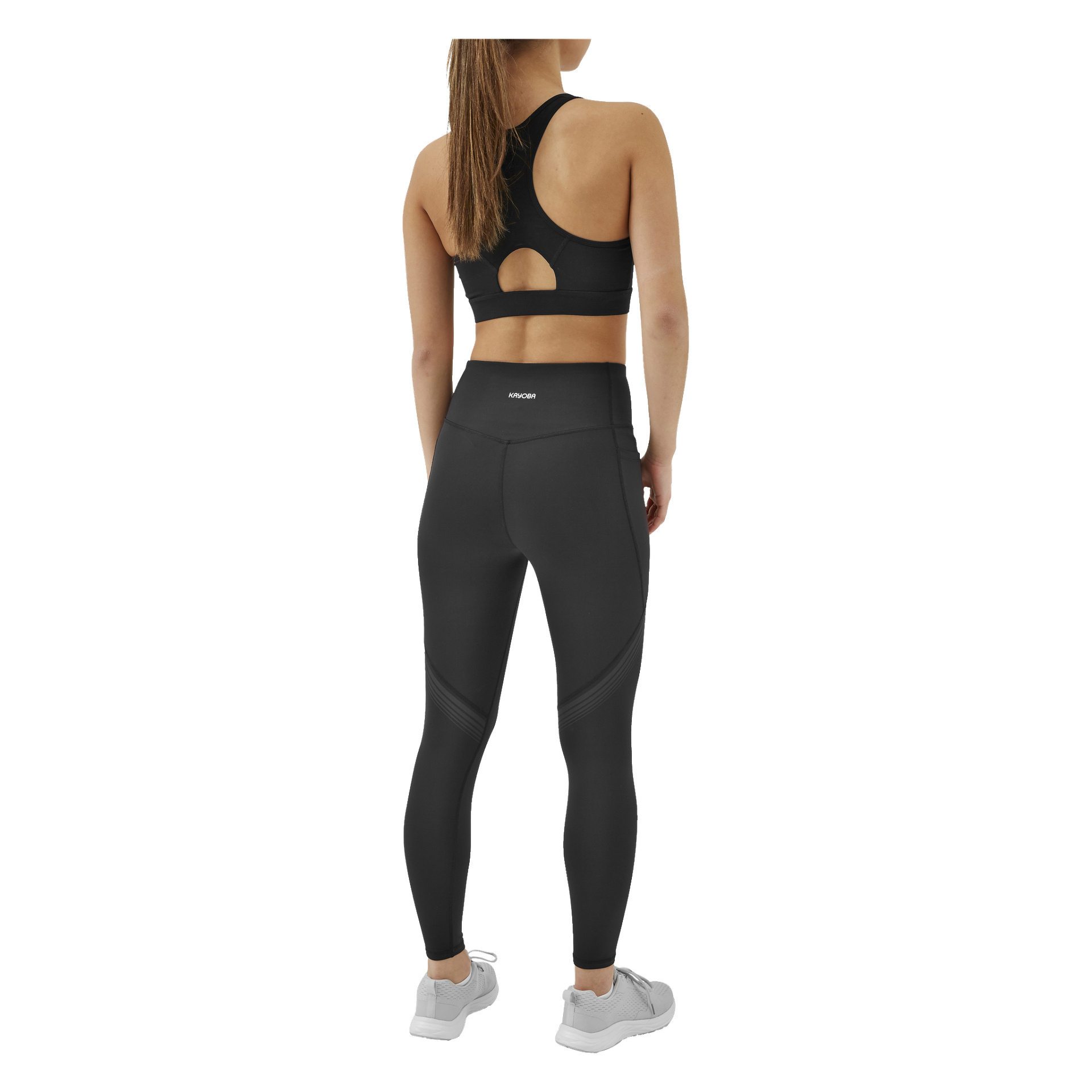 En kvinna med håret i hästsvans står med ryggen mot ryggen, klädd i svarta Kayoba Tights damleggings från Boston, en svart sport-bh och grå sportskor.