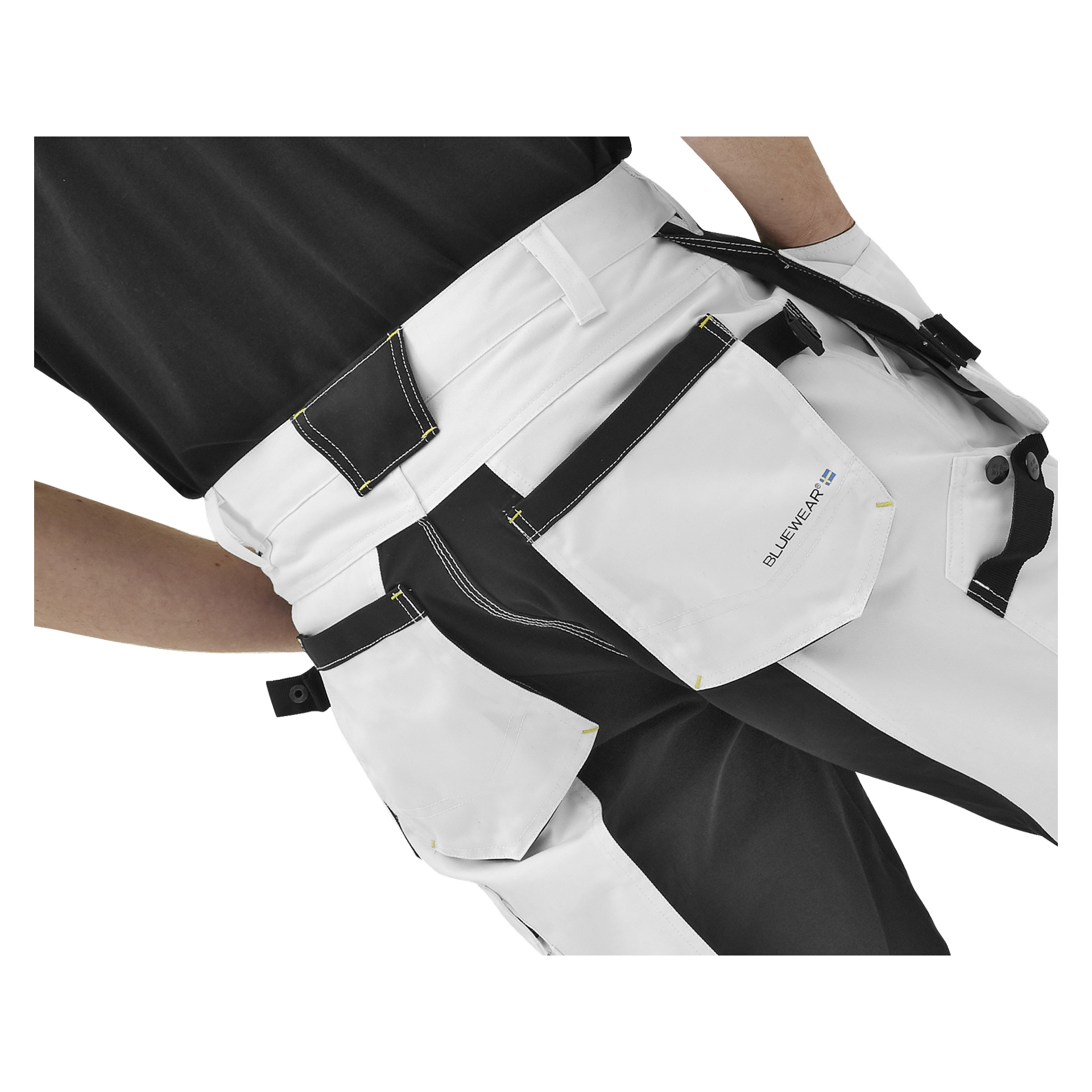 En person visas från midjan och nedåt bära Bluewear Men's Rex arbetsbyxor i vitt och svart, designade med flera fickor, gula sömmar och en synlig "Bluewear"-etikett.