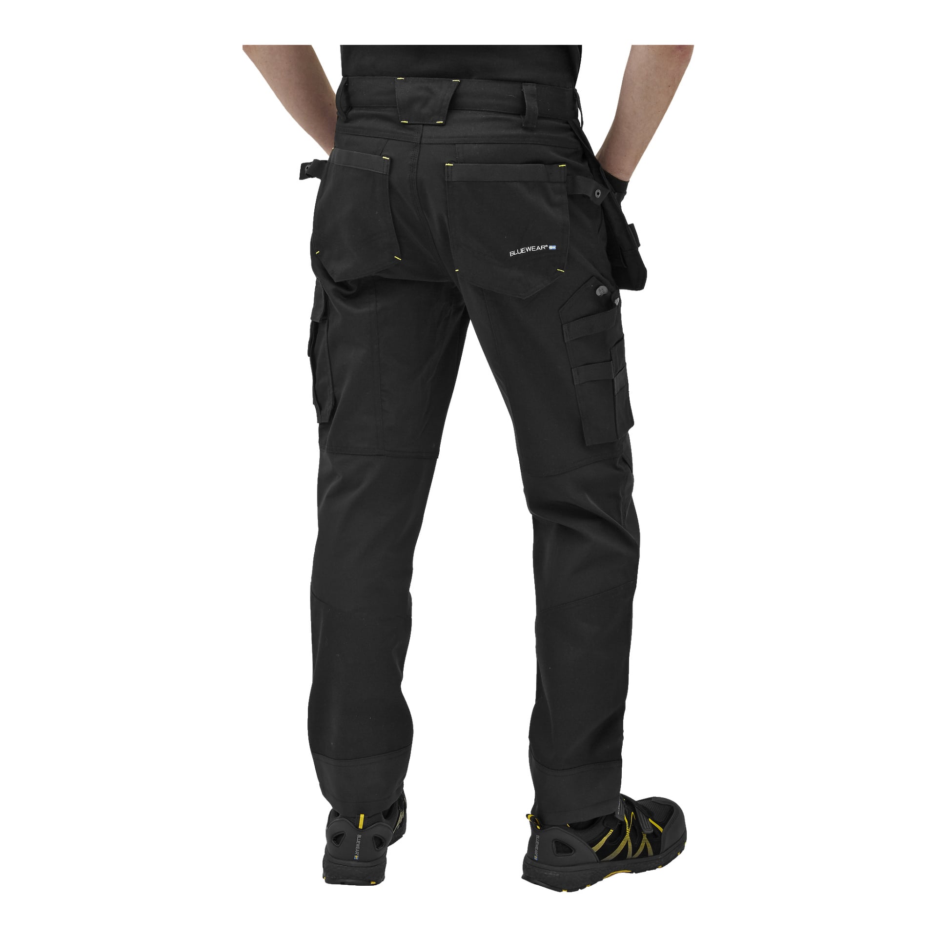 Sett bakifrån bär personen Bluewear Men's Rex svarta arbetsbyxor med flera fickor, framträdande gula sömmar, förstärkta knän och synlig märkning. De bär också svarta och gula arbetsskor. Personens överkropp och huvud är utanför bild.