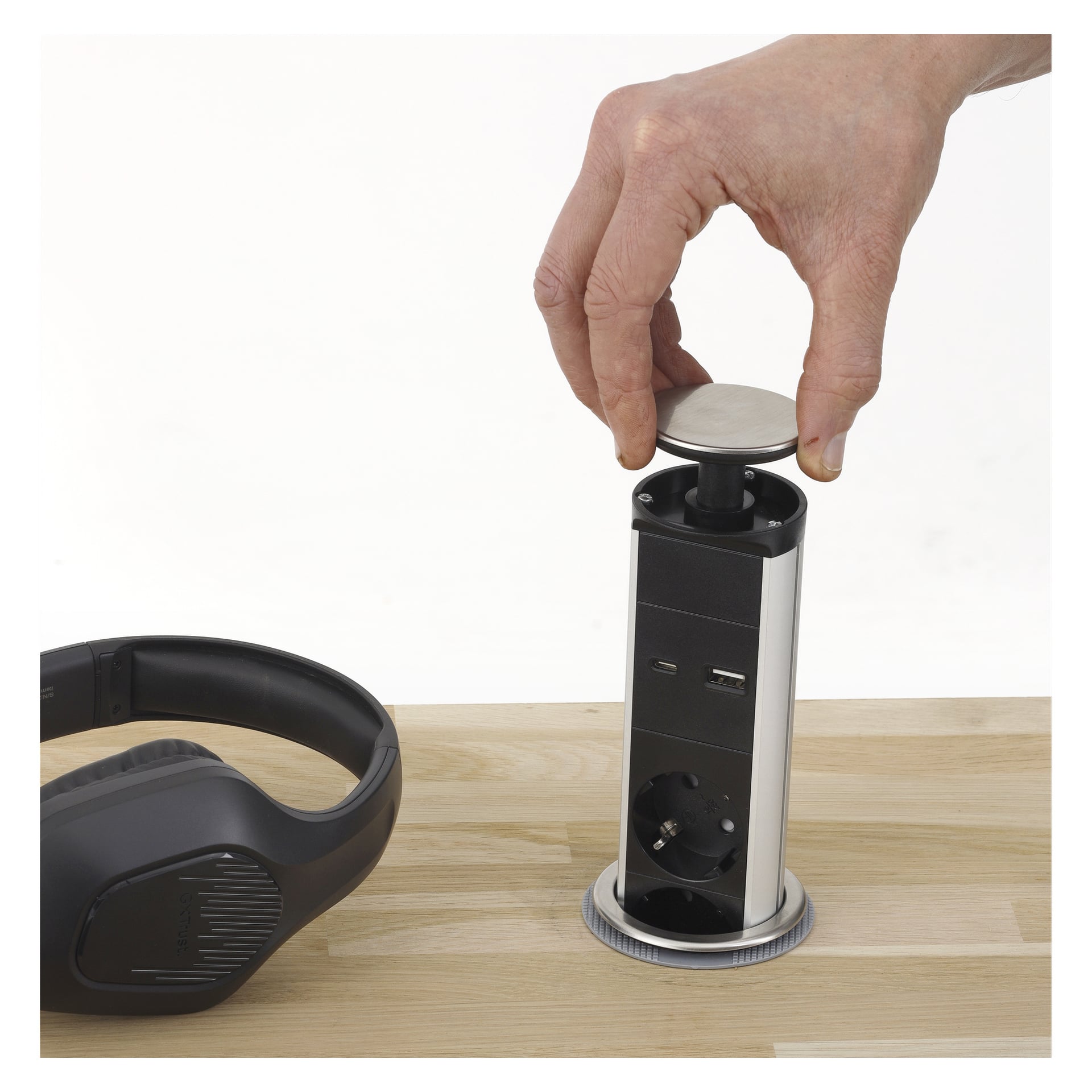 En hånd trykker ned på Ansluts Power Strip Pop-up, som er bygget inn i en treflate. Den uttrekkbare stikkontakten har USB-porter og en stikkontakt. Svarte hodetelefoner ligger på bordet i nærheten.