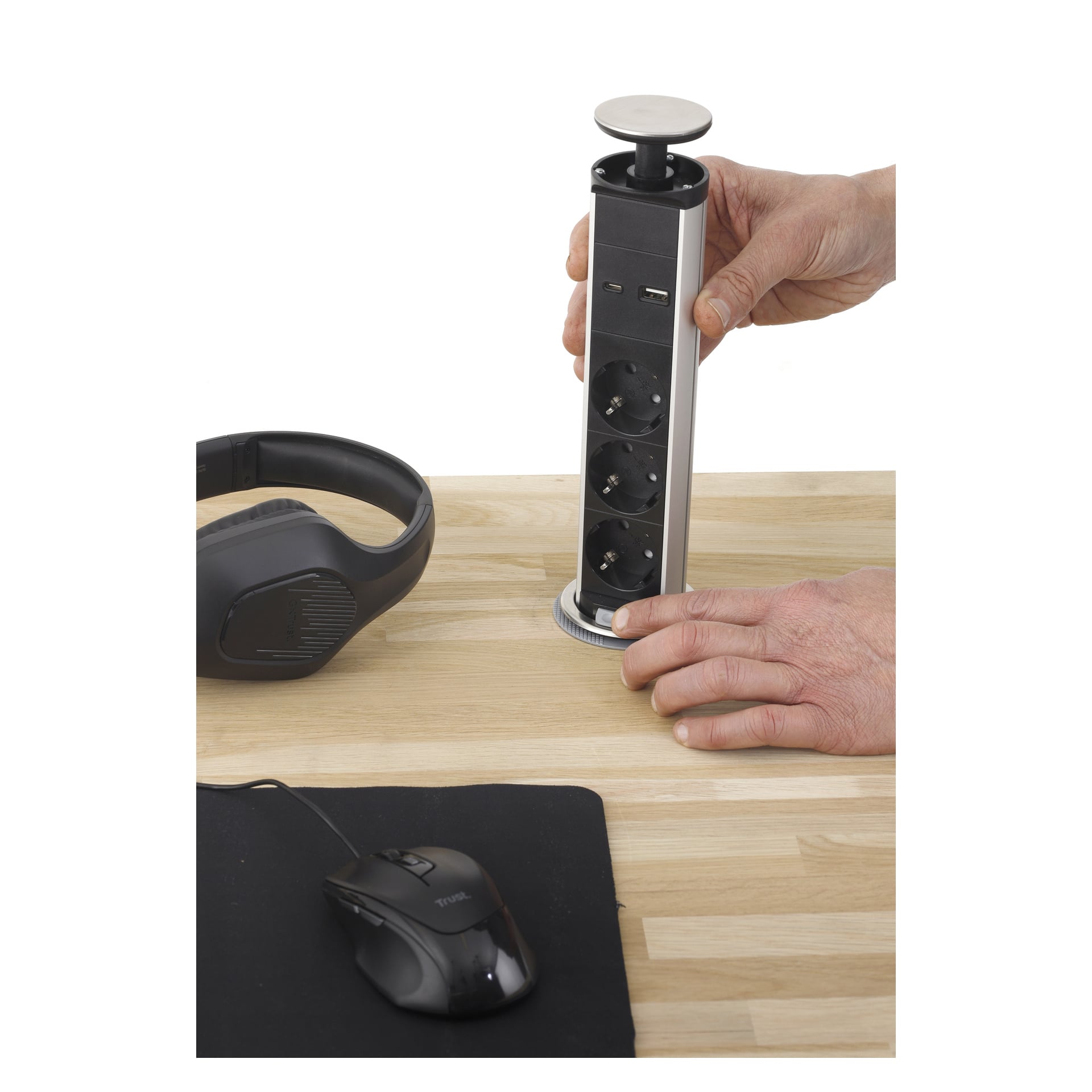 Noen løfter en Anslut Powerstrip Pop-up, med tre stikkontakter og to USB-porter, fra et skrivebord i tre som også inneholder et svart headset og en datamus.