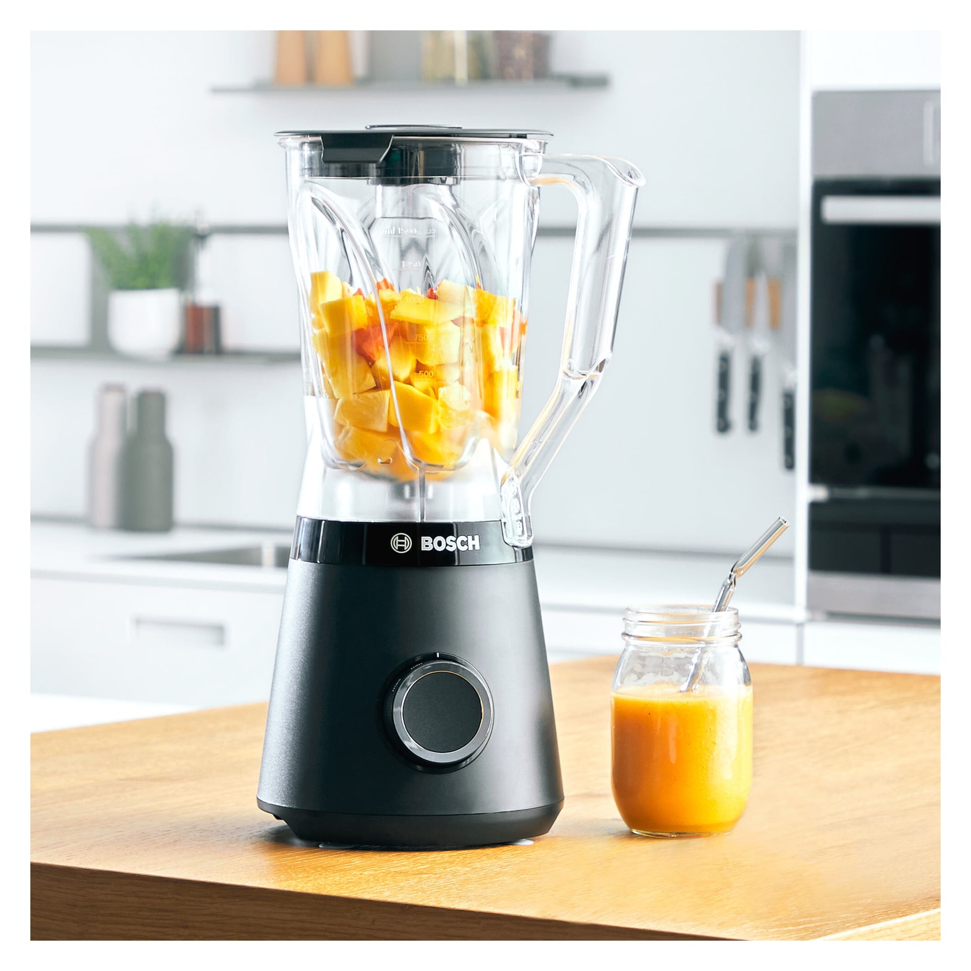 En Bosch Home Blender MMB6141B står på en köksbänk, fylld med bitar av apelsinfrukt. Bredvid står en glasburk med apelsinjuice och ett sugrör av metall. Bakgrunden visar väl upplysta kökshyllor och en diskhon.