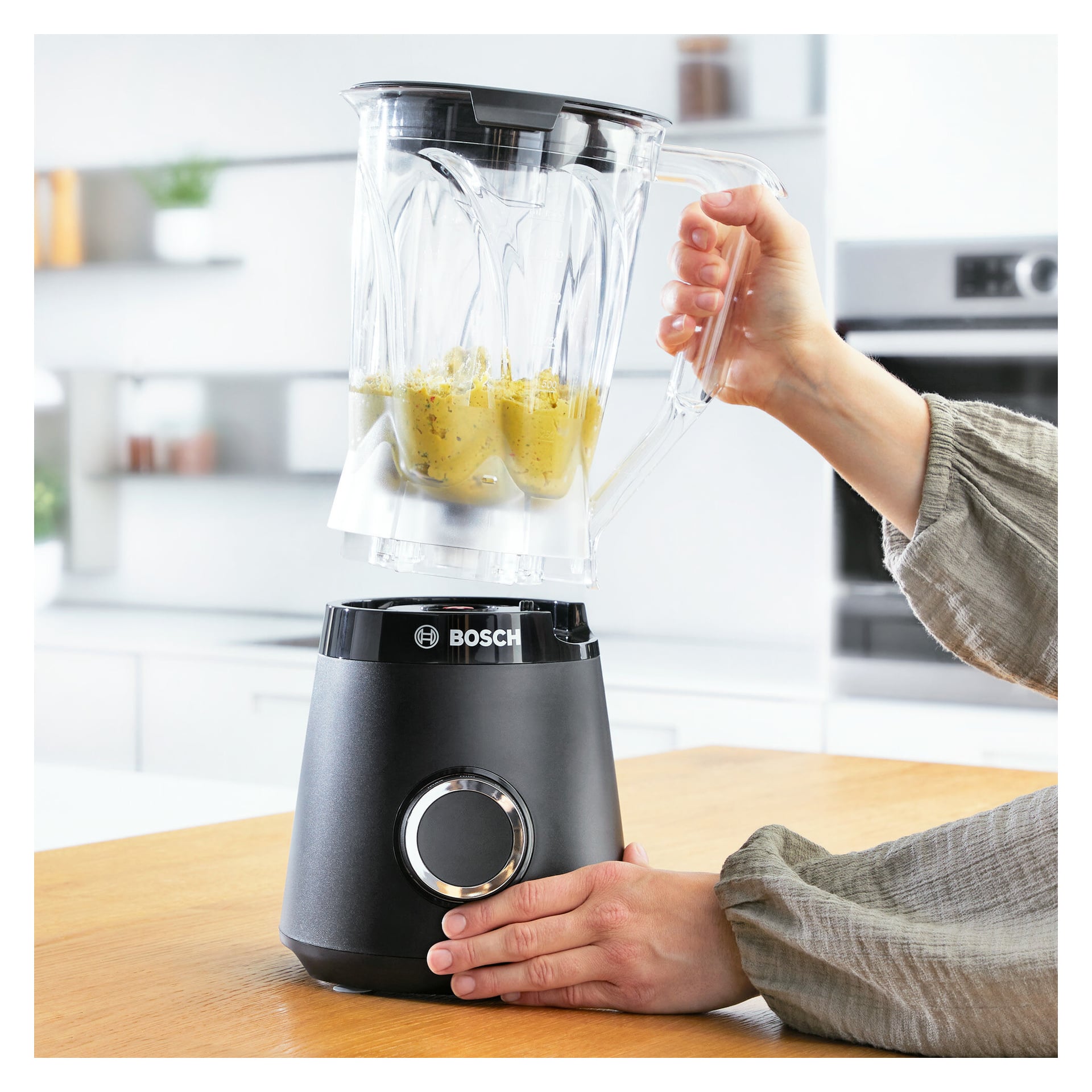 Någon trycker på en knapp på basen av en Bosch Home Blender MMB6141B, håller ner locket eftersom mixern innehåller en gul blandning, allt i en modern köksmiljö.
