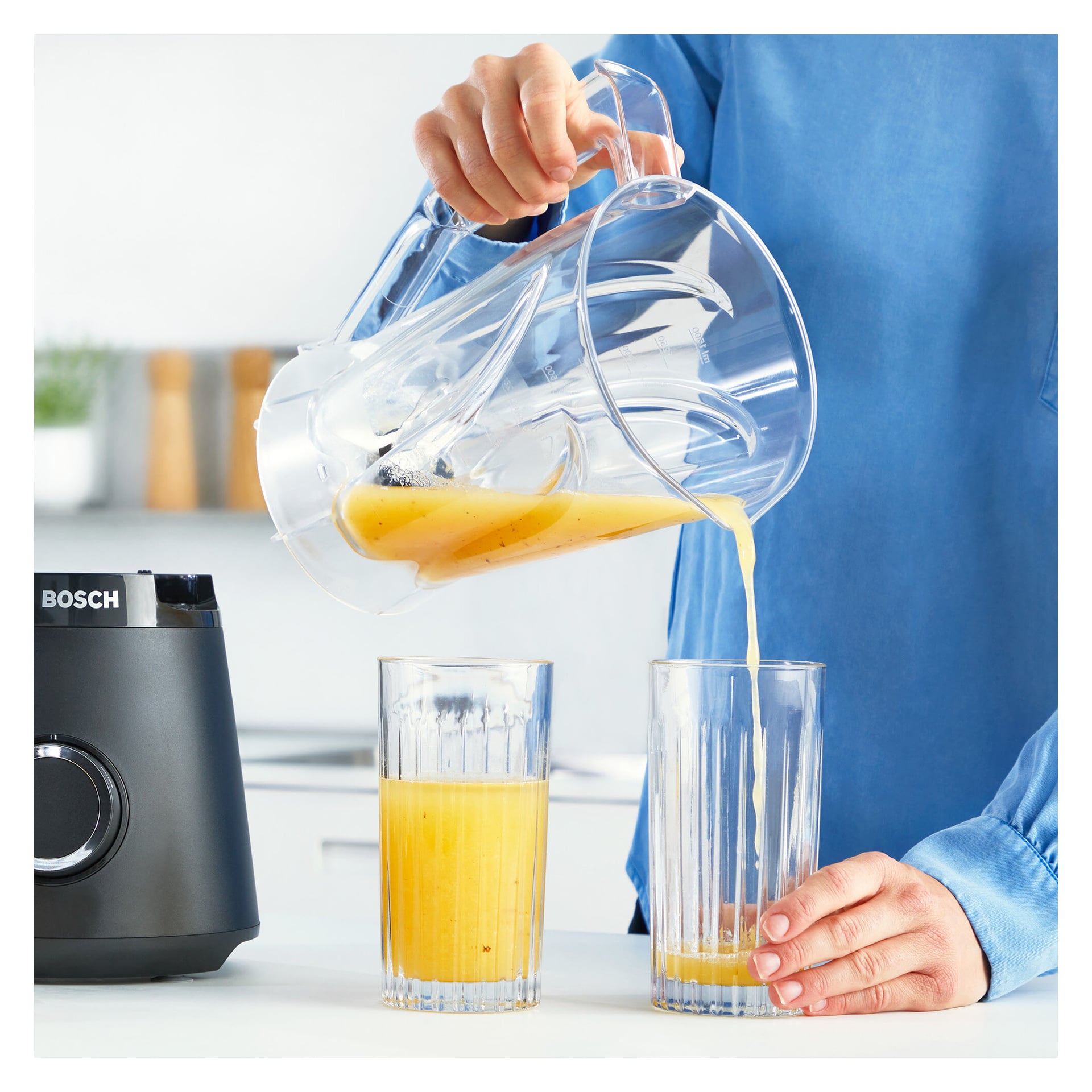 En person i blå skjorta häller apelsinjuice från Bosch Home Blender MMB6141B kanna i ett högt glas, med ett annat delvis fyllt glas bredvid. Mixerbasen finns till vänster, allt placerat i en köksmiljö.