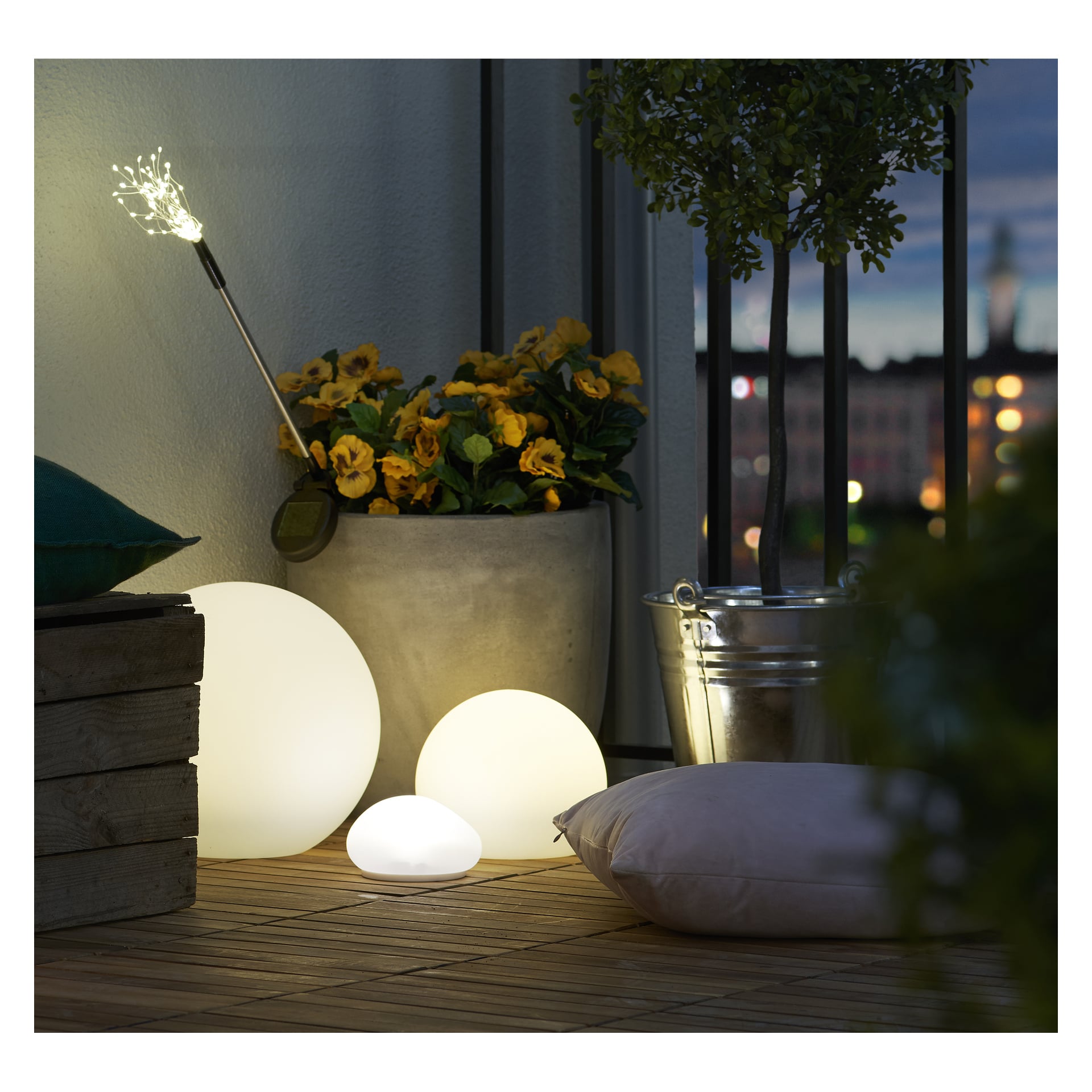 En varm balkong med en gul krukblomma, två upplysta Anslut LED-solglober, en liten rund lampa, en silverhink, en mjuk rosa kudde på en pallstol med en grön kudde ovanpå, och blinkande stadsljus synliga bortom.