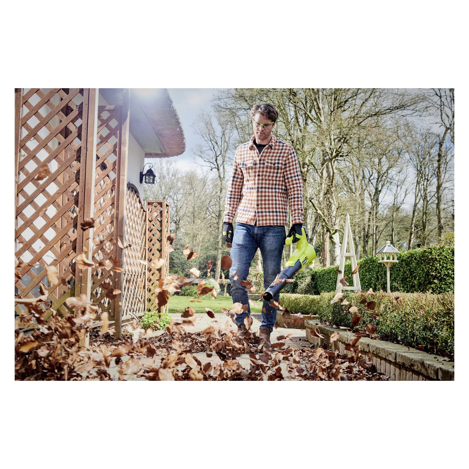 Mit Brille, Handschuhen und einem karierten Hemd entfernt man braune Blätter von einem Gartenweg neben einem Haus mit dem batteriebetriebenen Laubbläser Ryobi One+ RY18BLA-120. Bäume und Hecken säumen den Hintergrund.