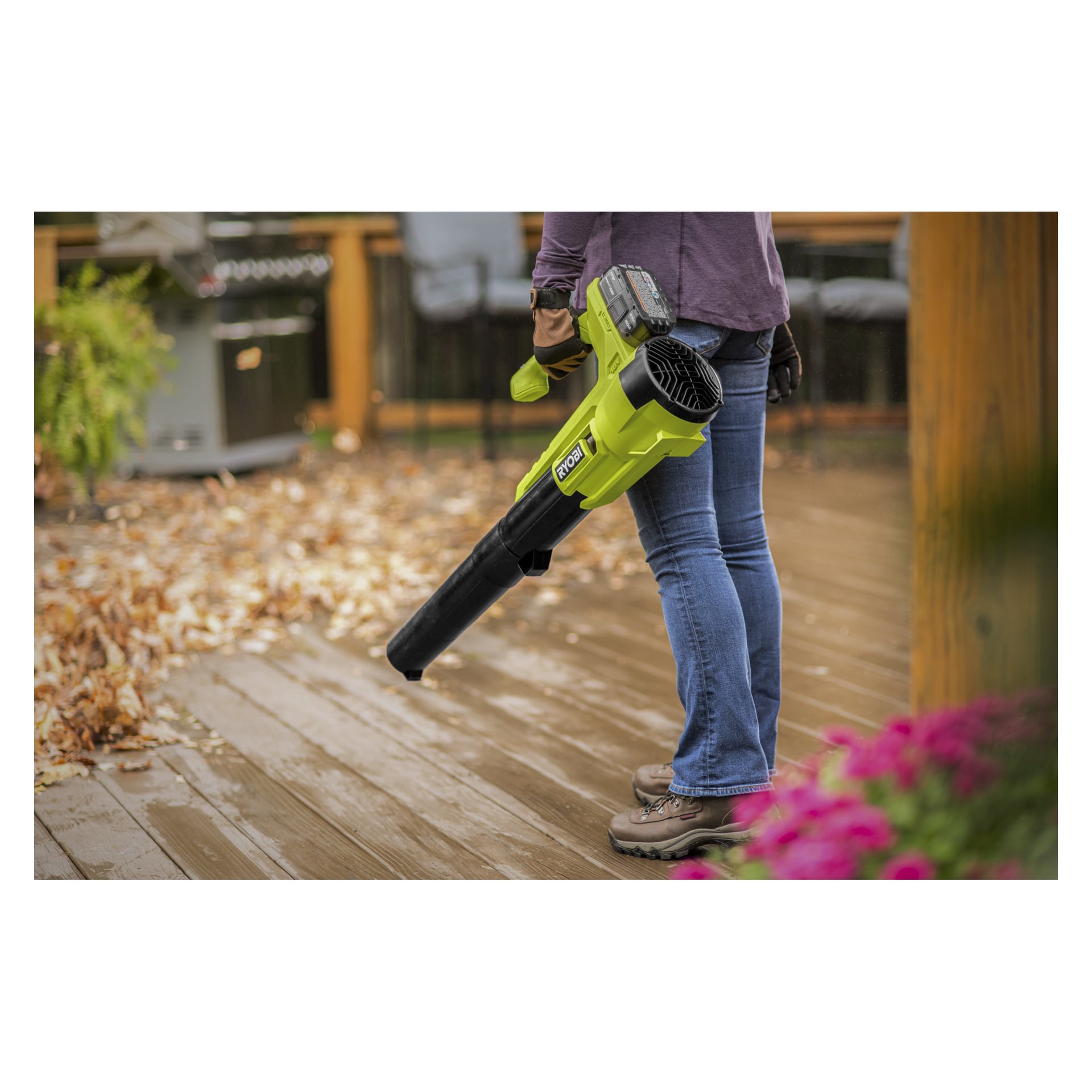 In Jeans und Stiefeln bedient man einen grünen Ryobi One+ RY18BLA-120 batteriebetriebenen Laubbläser, um verstreute Blätter von einer Holzterrasse zu entfernen, mit rosa Blumen und Gartenmöbeln im Hintergrund.