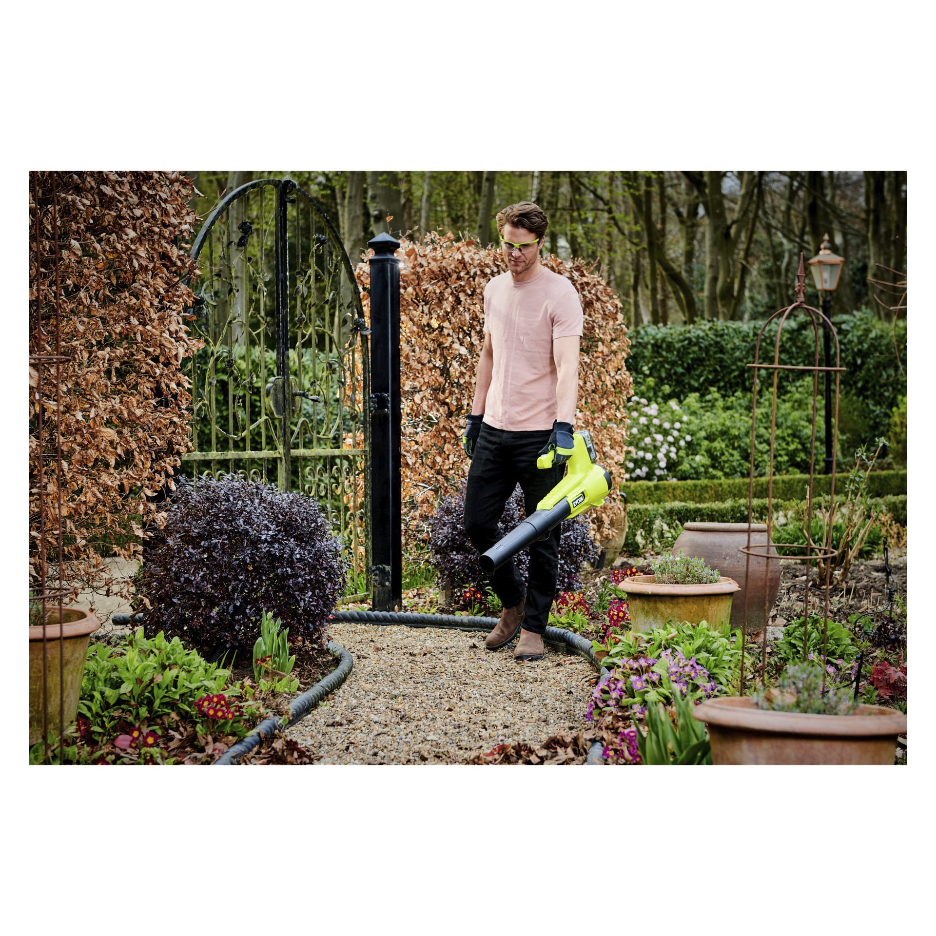 Ein Mann mit rosa Hemd, Handschuhen und Schutzbrille bedient einen Ryobi One+ batteriebetriebenen Laubbläser (Modell RY18BLA-120) und räumt Blätter von einem Kiesweg, der von üppigen Pflanzen und Topfblumen umgeben ist. Bäume und ein Metalltor sind hinter ihm sichtbar.