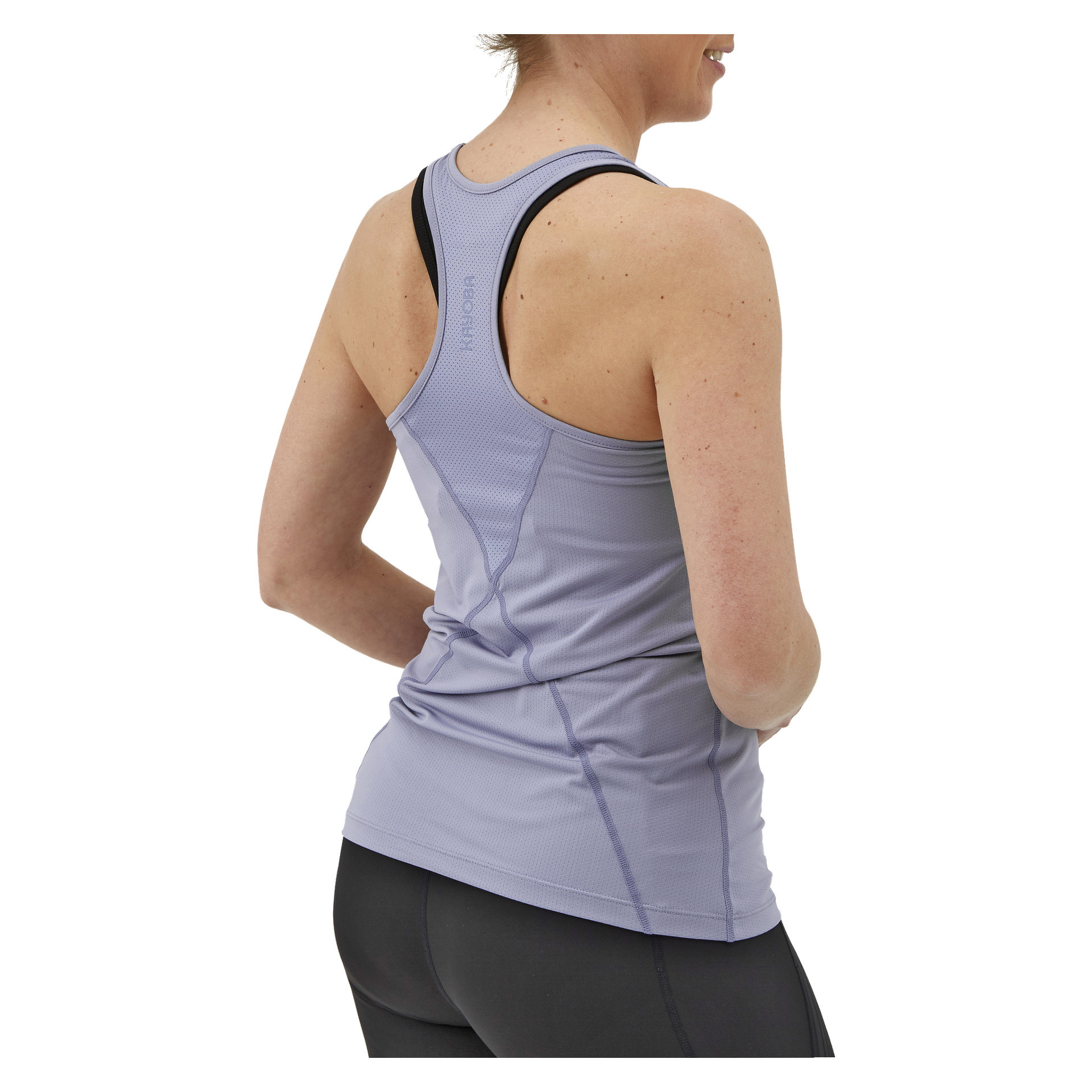 En kvinna står med ryggen mot kameran, iklädd en lavendel Kayoba Women's Training Tank Top Albany och svarta leggings. Racerback-designen visar framträdande sömdetaljer och en logotyp nära halsringningen. Hennes hår är uppsatt, och hon har händerna vilande på höfterna.