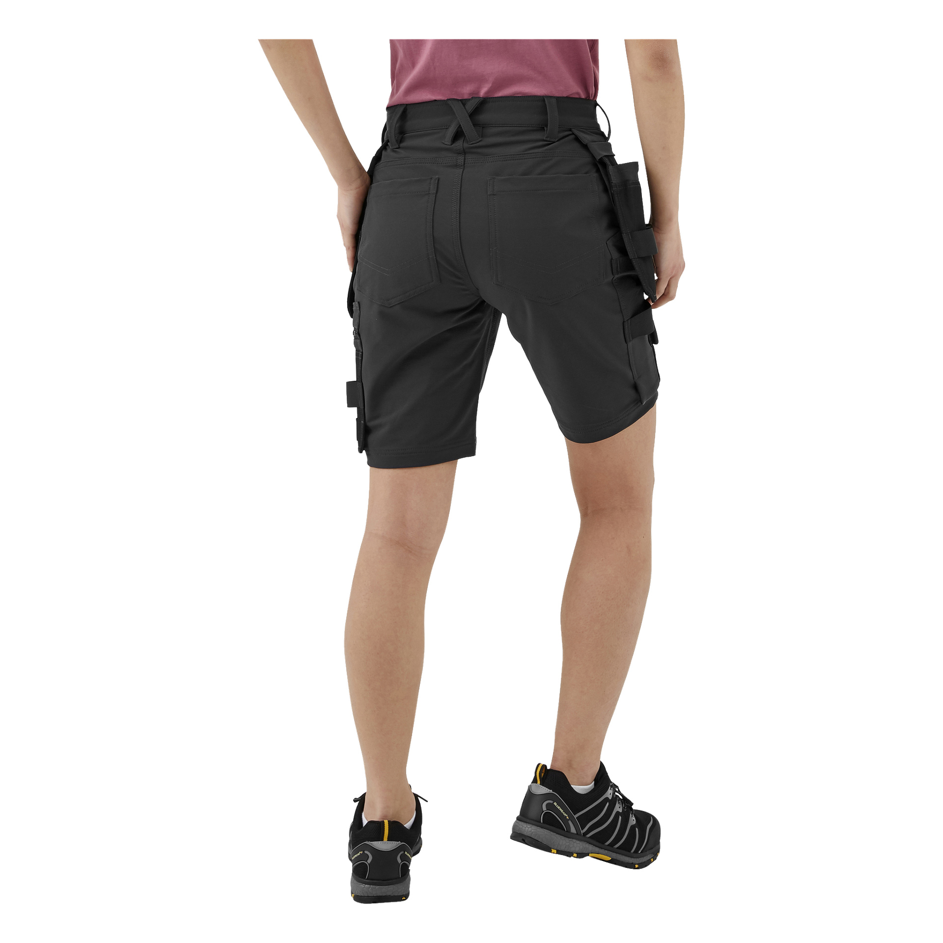 Od tyłu widziana jest osoba ubrana w czarne robocze spodenki robocze Bluewear Pro Workwear Women's Stella z bocznymi kieszeniami, fioletową koszulę oraz czarne buty sportowe z żółtymi akcentami.