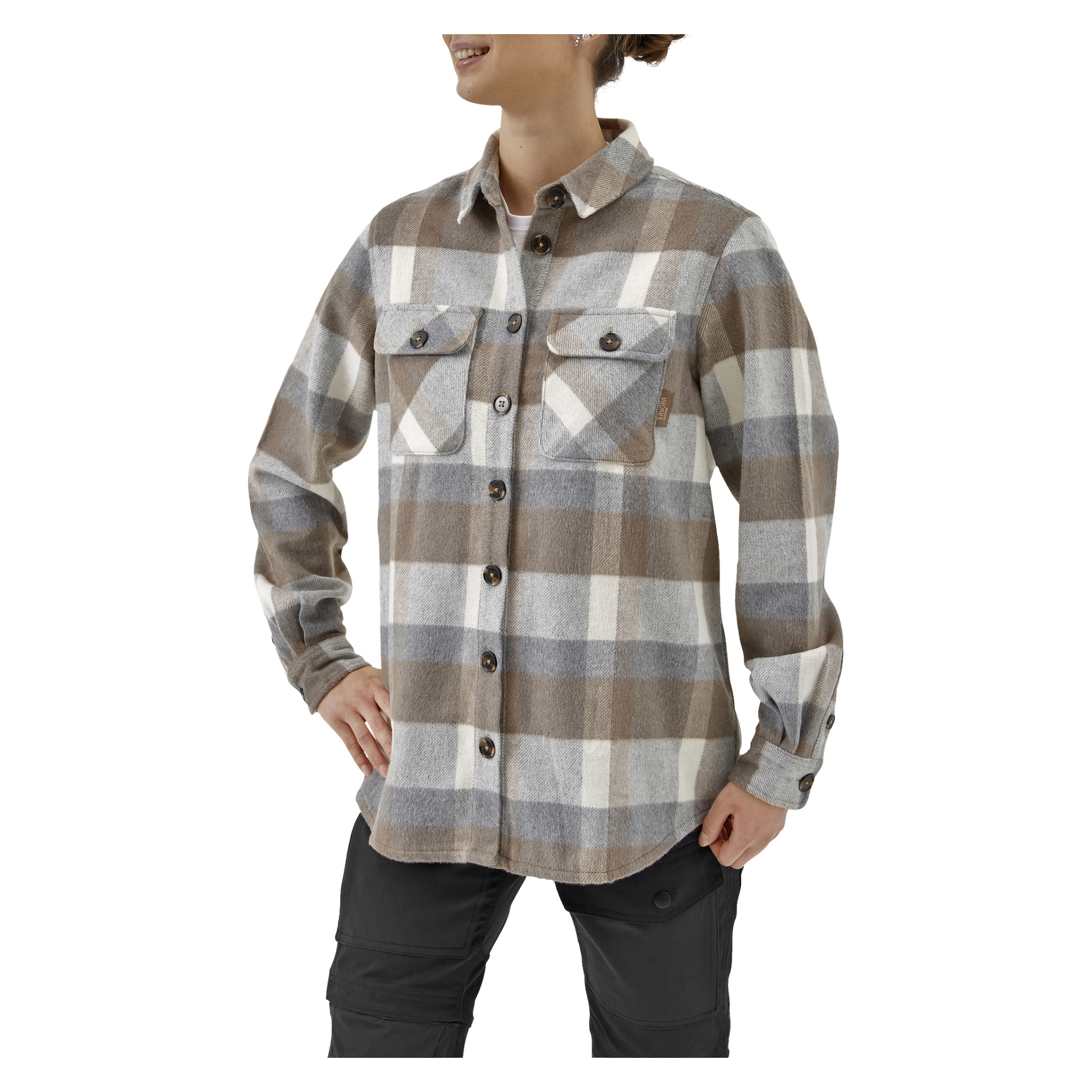 Noen modellerer Kayoba Outdoor Women's overdress Idhult, en rutete skjorte i brunt, grått og hvitt med to brystlommer. De kombinerer det med mørke bukser og poserer med en hånd på hoften, smilende.