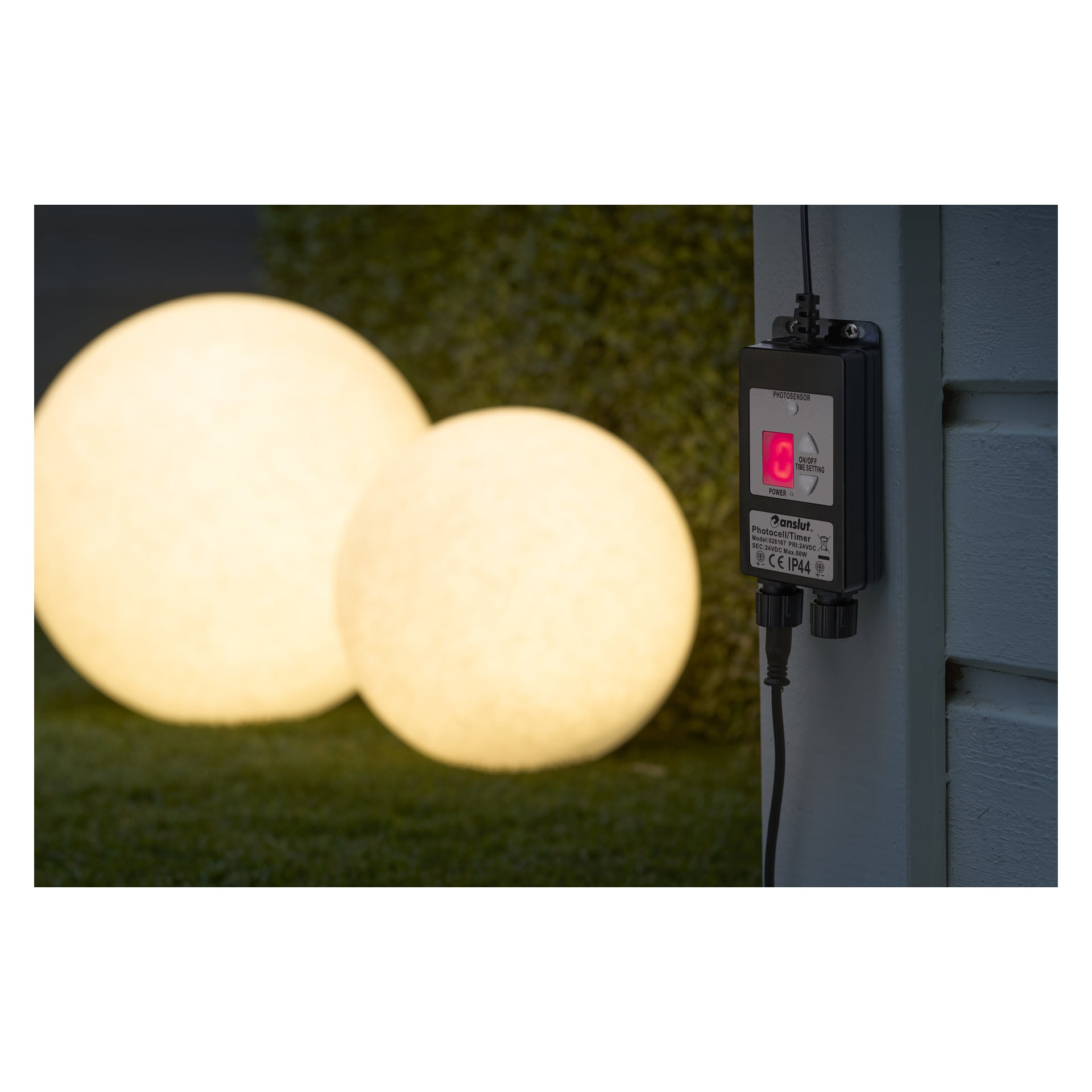 Et svart Anslut Twilight-sensor Connectable System med rød strømindikator er festet til en lysvegg. Bak den sitter to store glødende kuler på gresset ved en hekk.