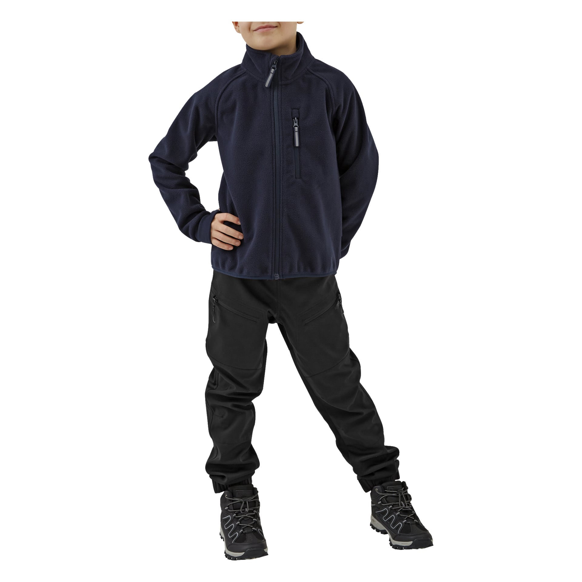 Ein Kind steht mit einer Hand in der Hüfte, gekleidet in schwarze Kinderhosen von Kayoba Outdoor mit Reißverschlusstaschen, einer dunklen Reißverschlussjacke und dunklen Wanderschuhen. Das Gesicht des Kindes ist teilweise sichtbar.
