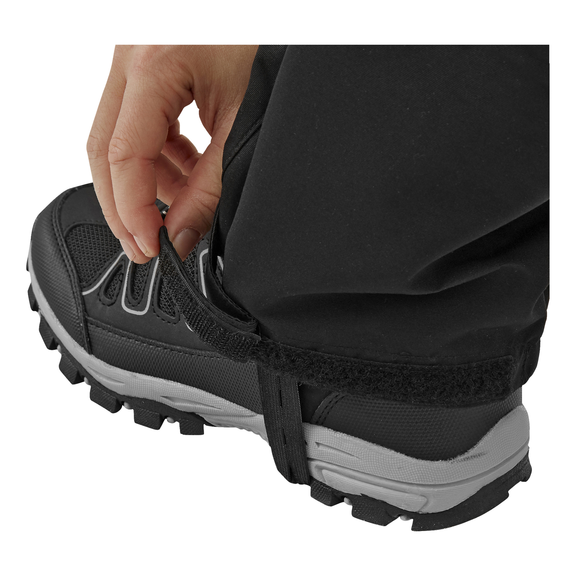 Eine Hand befestigt einen Velcro-Riemen über der Ferse eines schwarzen Wanderstiefels mit robuster Sohle und grauen Akzenten und zieht ihn um den Saum der schwarzen Kayoba Outdoor Kinderhosen fest.