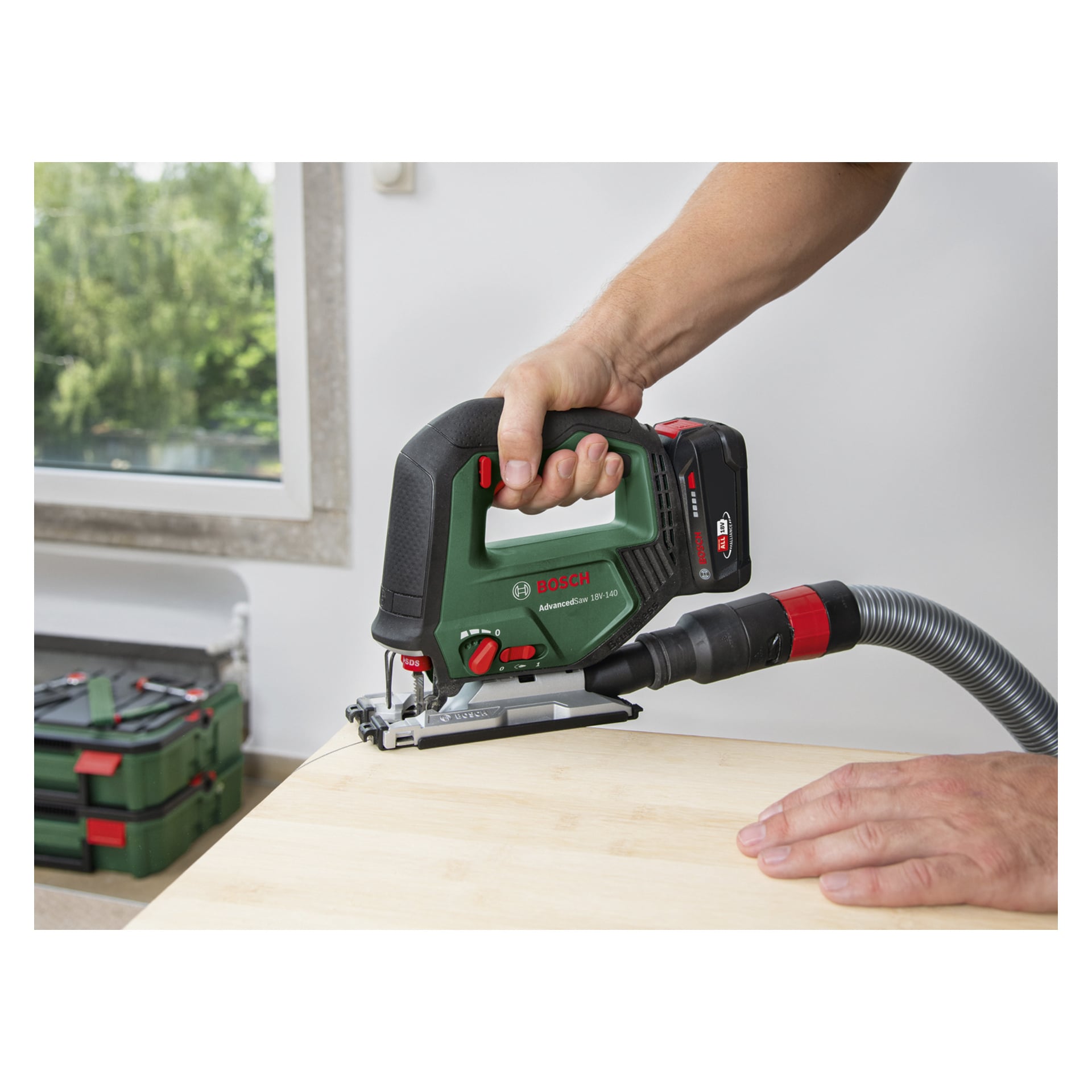 Noen bruker en Bosch Power 4 All AdvancedSaw 18V-140 batteri-stikksag med støvavtrekker, og kutter kanten på en lett treplate. I bakgrunnen er grønne verktøykasser synlige, og et vindu viser trær utenfor.