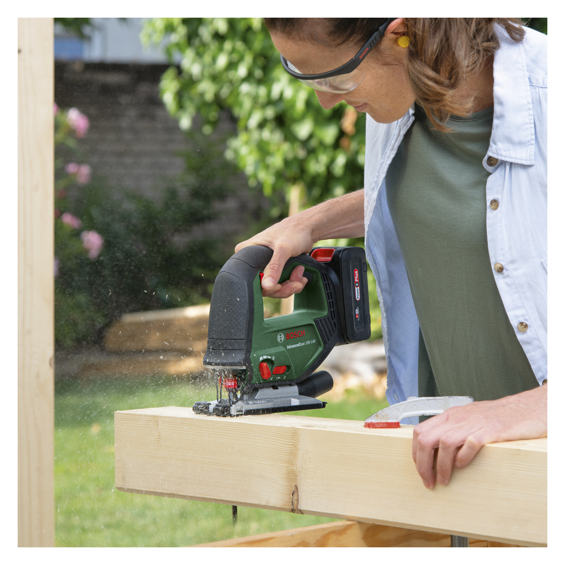 En person med vernebriller skjærer en trebjelke ute med et Bosch Power 4 All Battery-drevet puslespill AdvancedSaw 18V-140, og skaper en sky av trestøv mens livlig grønt omgir scenen.