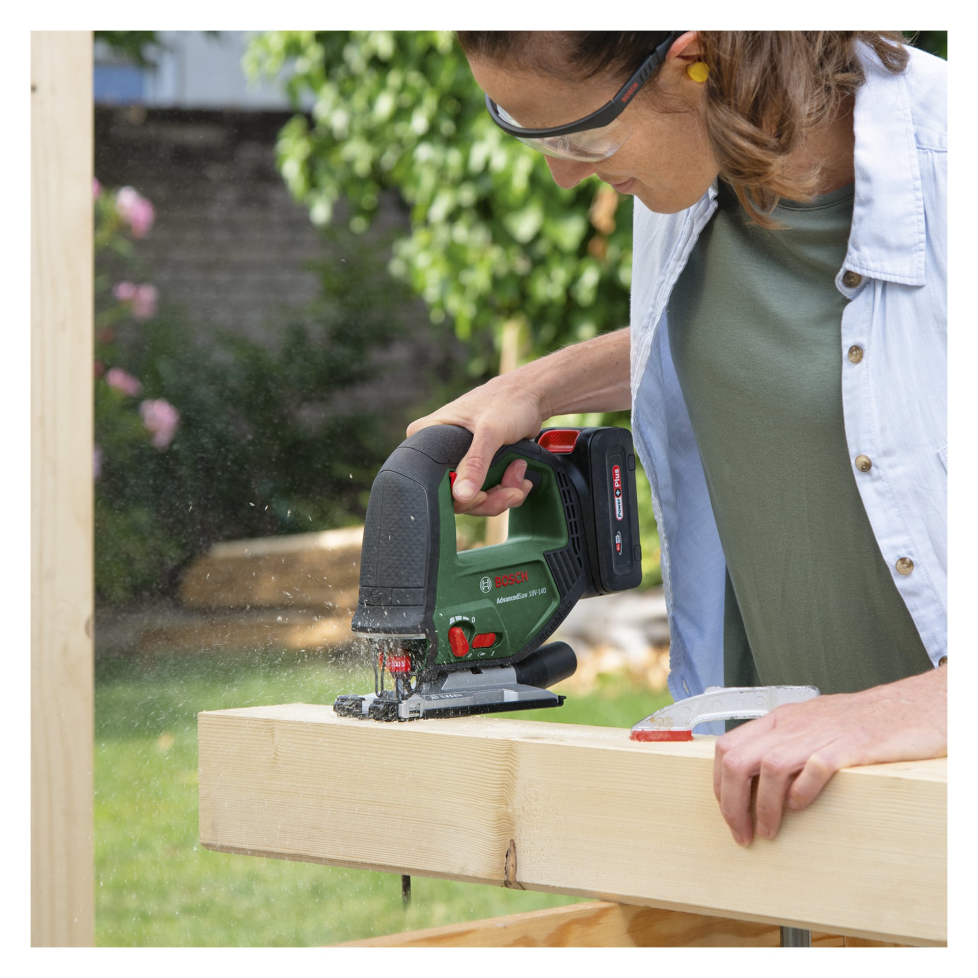 En person med vernebriller skjærer en trebjelke ute med et Bosch Power 4 All Battery-drevet puslespill AdvancedSaw 18V-140, og skaper en sky av trestøv mens livlig grønt omgir scenen.