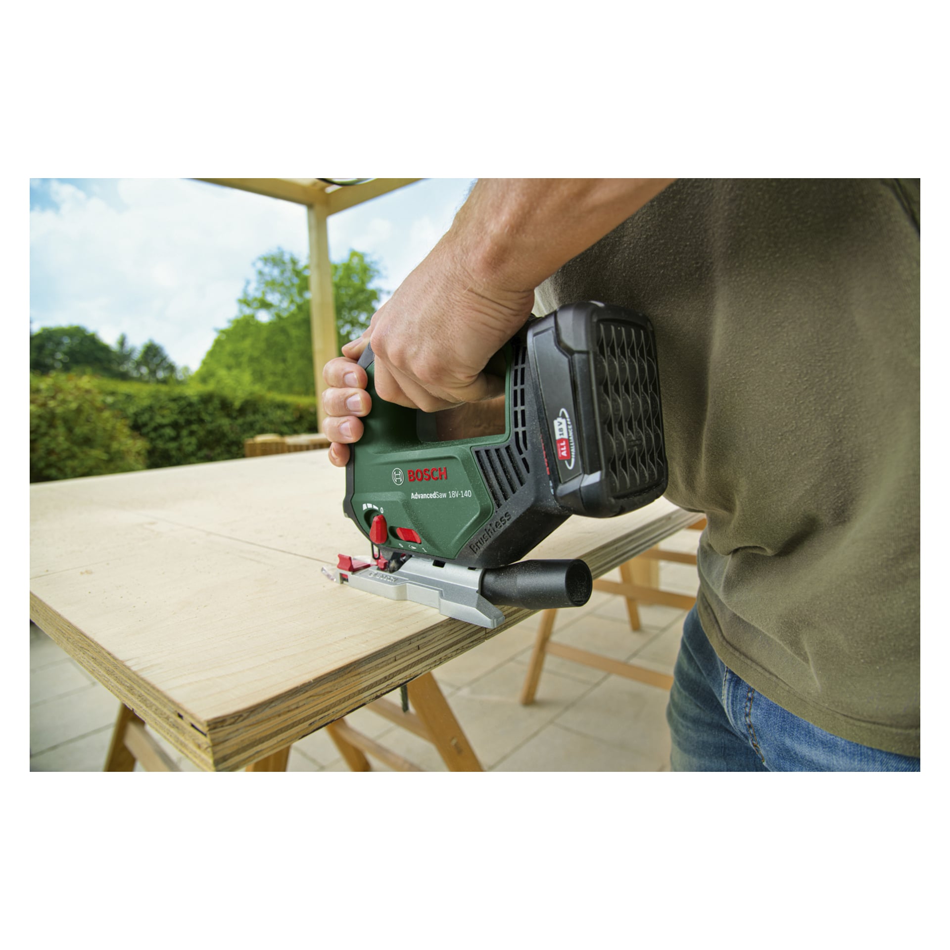 Noen bruker et Bosch Power 4 All AdvancedSaw 18V-140 batteri-stikksag for å kutte kryssfiner på en utendørs arbeidsbenk. Hånden deres holder verktøyet sikkert, følger en merket retningslinje, med løvtrær og planter i bakgrunnen.