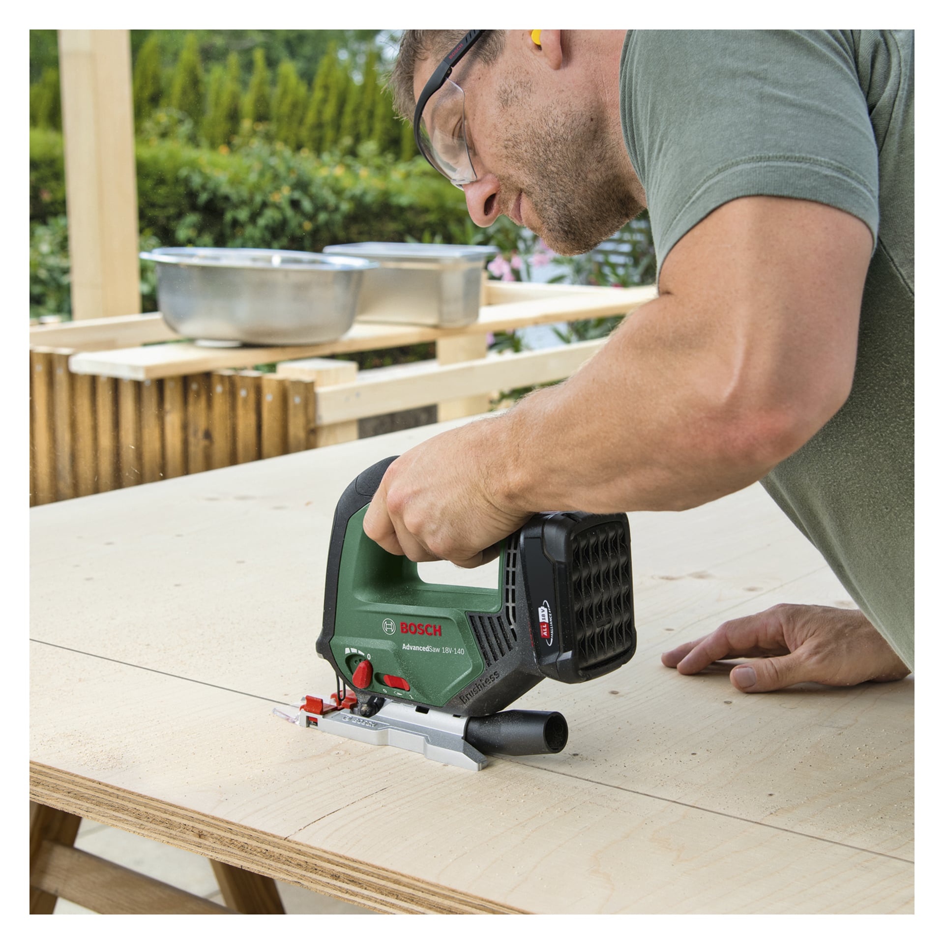 En person med vernebriller betjener en Bosch Power 4 All Battery stikksag AdvancedSaw 18V-140, og skjærer en rett linje i kryssfiner på en utendørs arbeidsbenk.