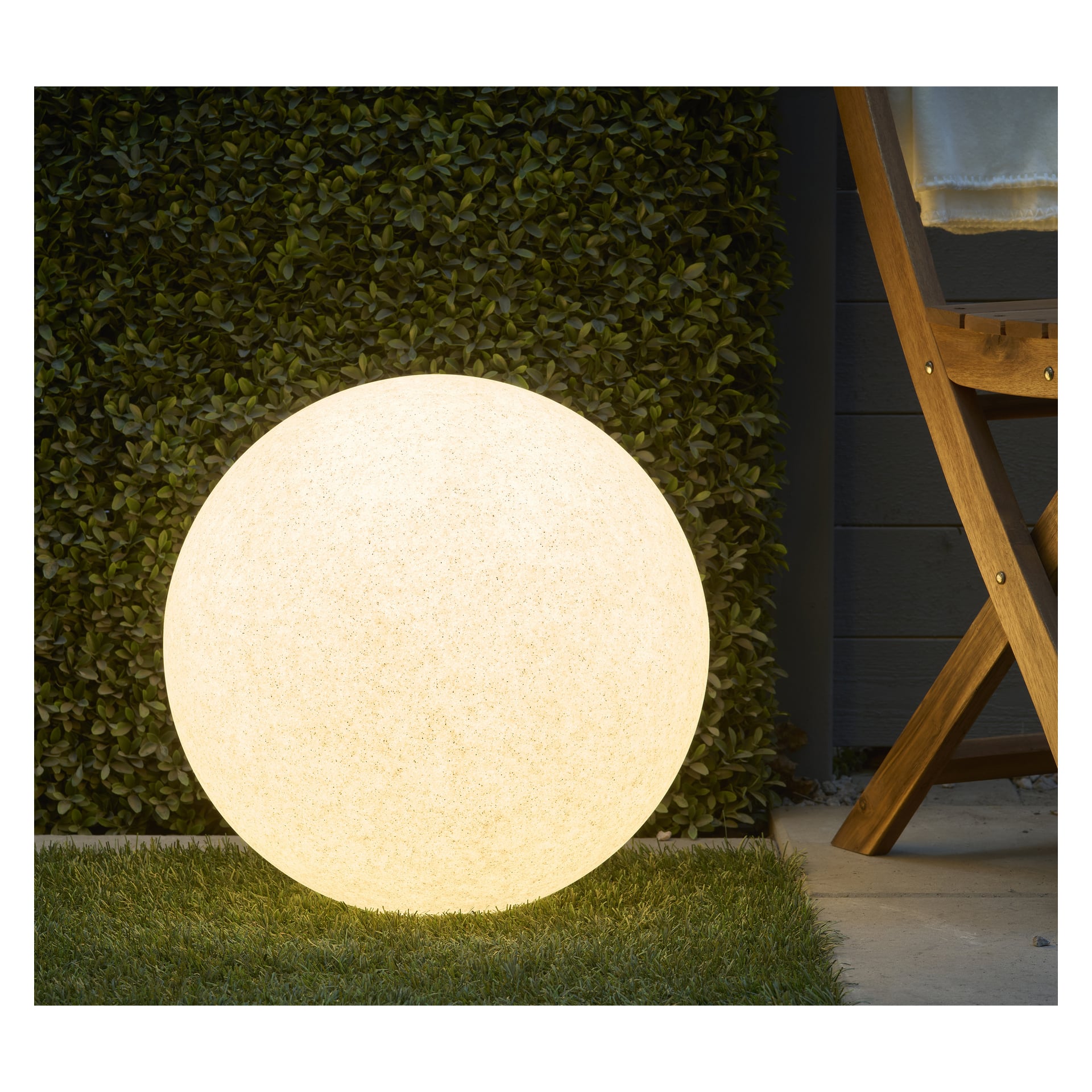 En glødende Anslut LED-kule fra Connectable System står på gress nær en pent trimmet hekk og ved siden av en del av en sammenleggbar trestol. Lampen bader scenen i varmt lys og lyser opp utendørsmiljøet i skumring eller natt.