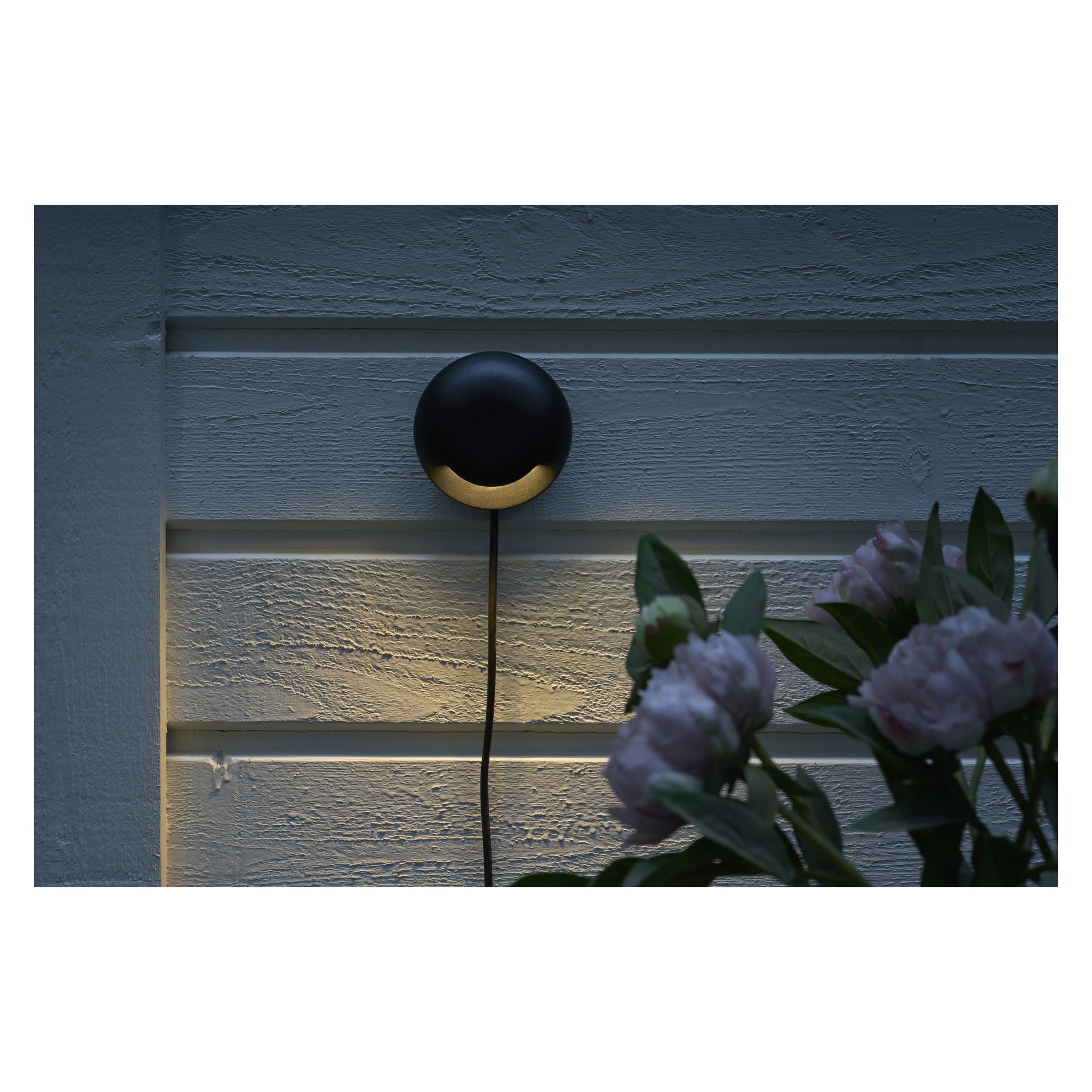 Pyöreä musta Anslut LED -seinävalo Connectable System -kokoelmasta on kiinnitetty vaakasuoriin puupaneeleihin, jotka heittävät pehmeän alaspäin suuntautuvan valon. Musta naru roikkuu valaisimesta, ja alakulmassa oikeassa etualalla näkyy sumeita vaaleanpunaisia kukkia vihreine lehdine.