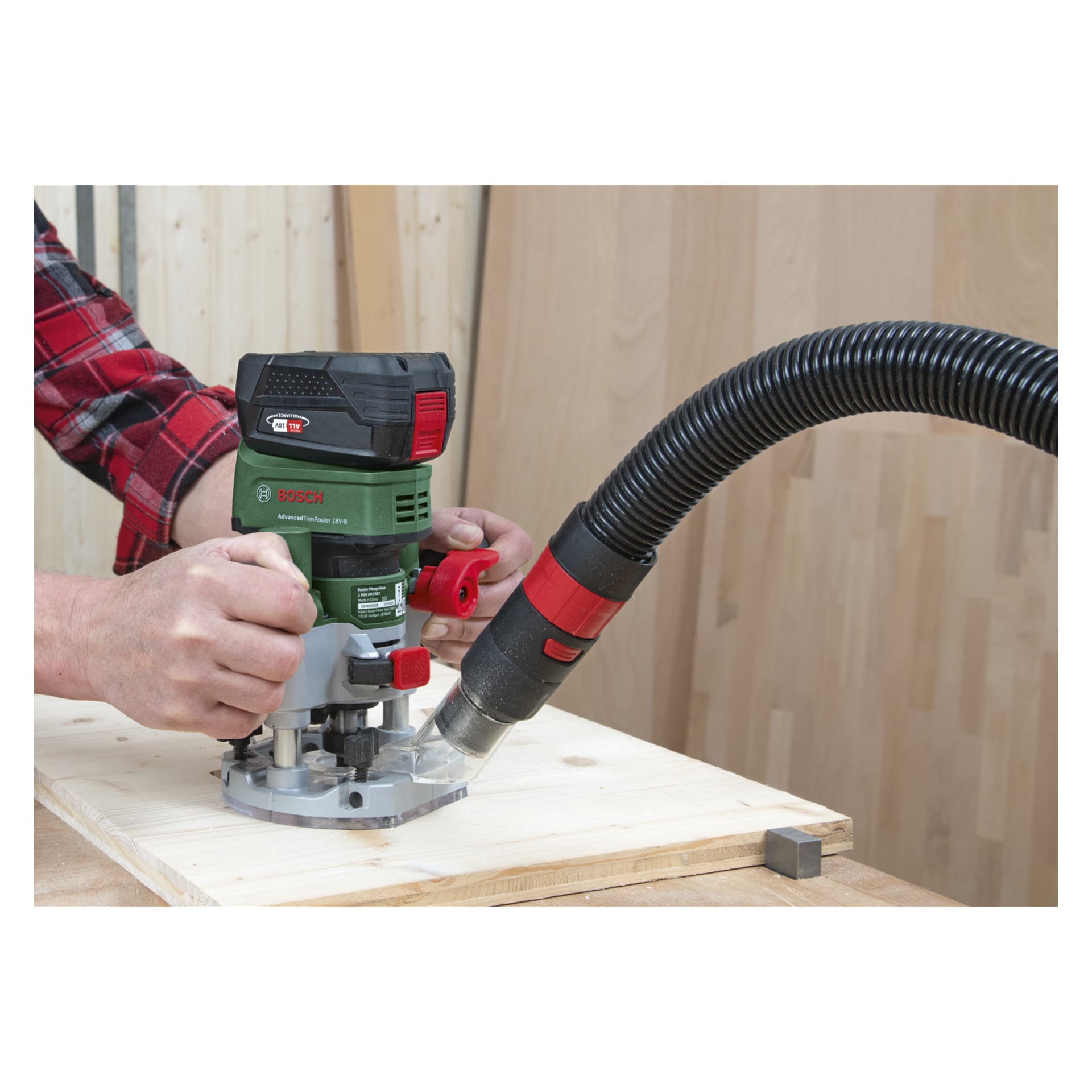 Jemand bedient den Bosch Power 4 All AdvancedTrimRouter 18V-8 mit einer Fräs-Tauchbasis auf einem Holzbrett. Ein Vakuumschlauch wird angeschlossen, um Sägemehl während der Holzbearbeitung aufzufangen.