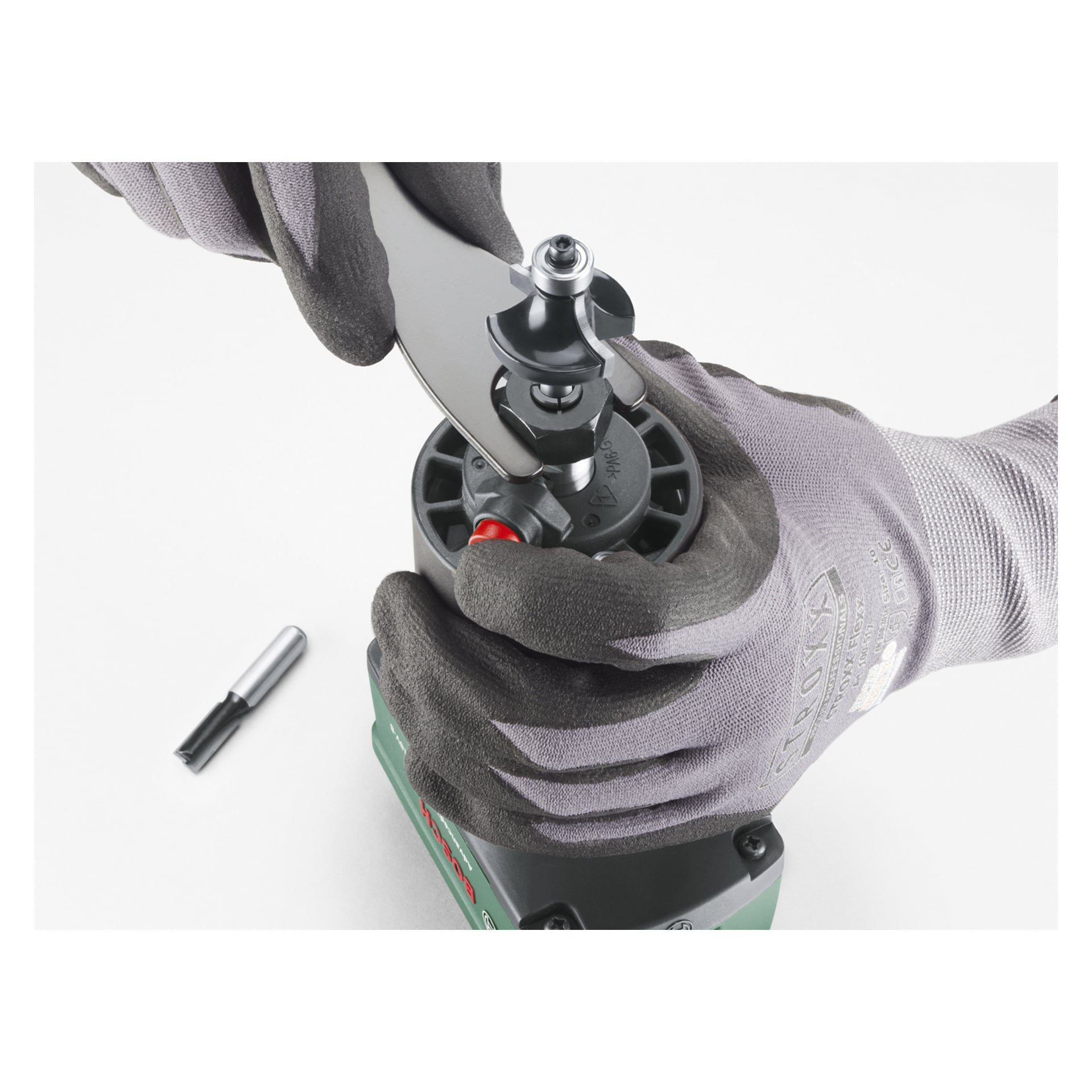 Grau behandschuhte Hände benutzen einen Schraubenschlüssel, um einen Fräser am Bosch Power 4 All Combination AdvancedTrimRouter 18V-8 mit Plunge-Basis zu befestigen; Ein weiterer Fräser liegt in der Nähe.