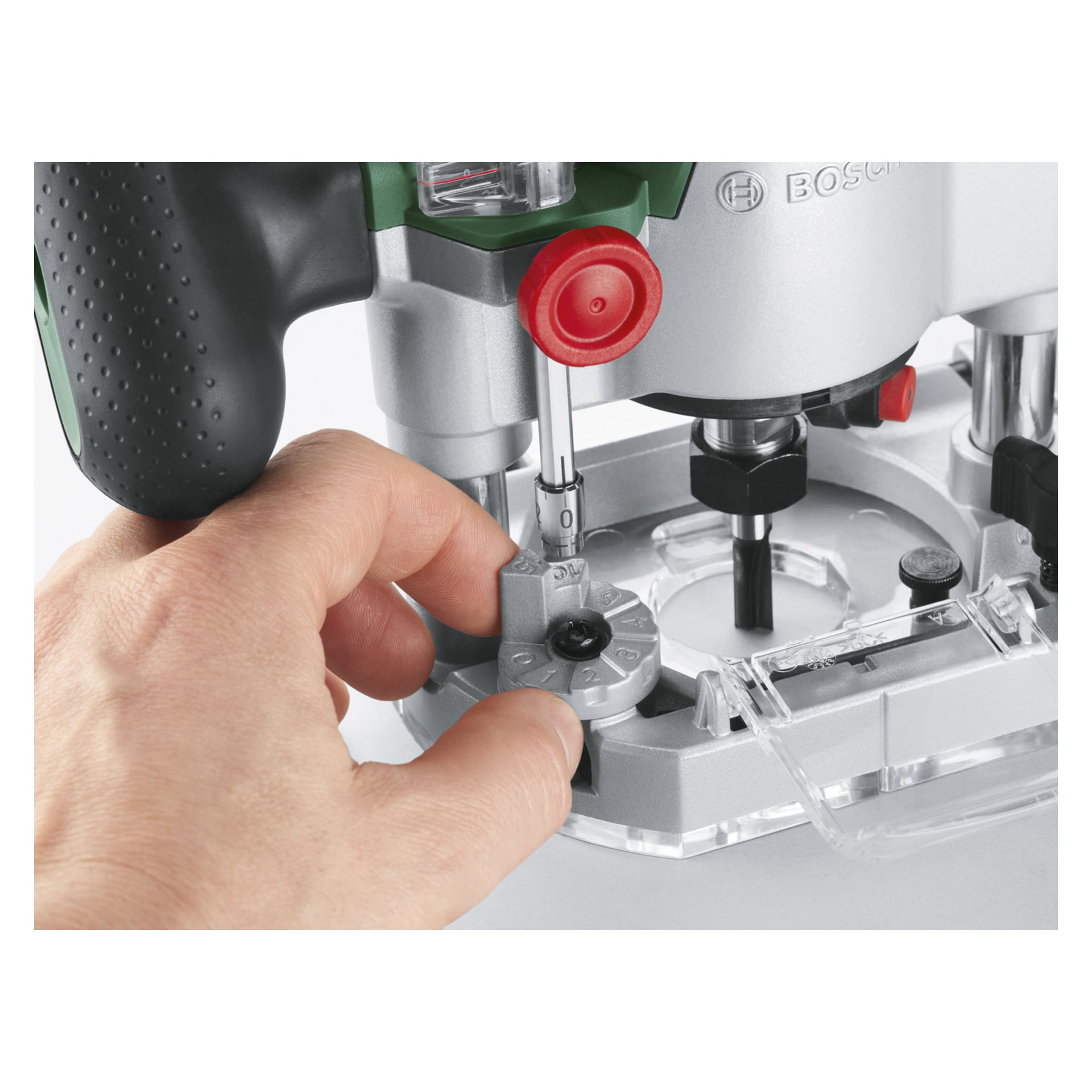 Eine Hand verändert eine Steuerung am Bosch Power 4 All AdvancedTrimRouter 18V-8 mit Plunge Base und hebt ein graues Zifferblatt und einen markanten roten Knauf neben einem durchsichtigen Kunststoffschutz hervor. Das Werkzeug sieht makellos aus und wird vor einem unauffälligen Hintergrund fotografiert.