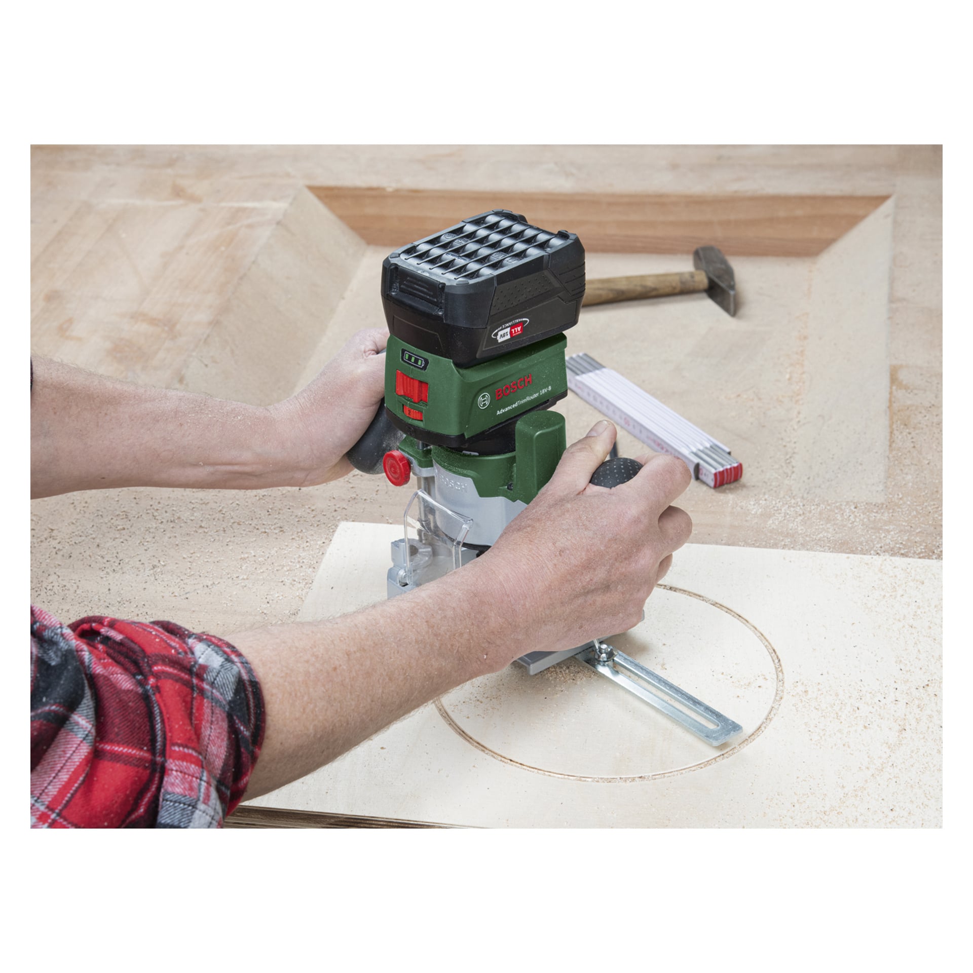 Eine Person bedient einen grün-schwarzen Bosch Power 4 All AdvancedTrimRouter 18V-8 mit Fräs-Tauchbasis, wobei eine kreisförmige Umrisse ins Holz geschnitten wird; Im Hintergrund der Werkbank sind ein Hammer und ein gefaltetes Lineal zu sehen.