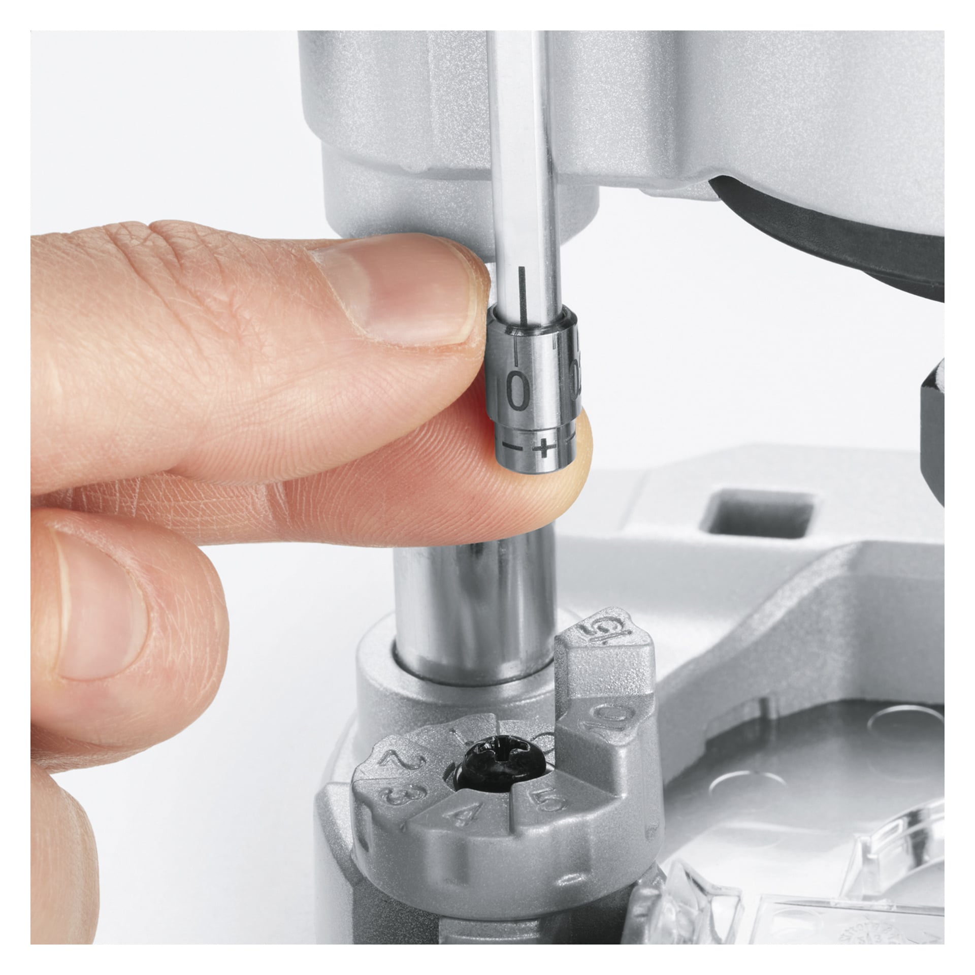 Ein Zeiger dreht den nummerierten Knopf, der derzeit auf Null gestellt ist, des Bosch Power 4 All AdvancedTrimRouter 18V-8 mit Plunge Base. Das markierte Zifferblatt des Werkzeugs und verschiedene mechanische Teile sind deutlich sichtbar.