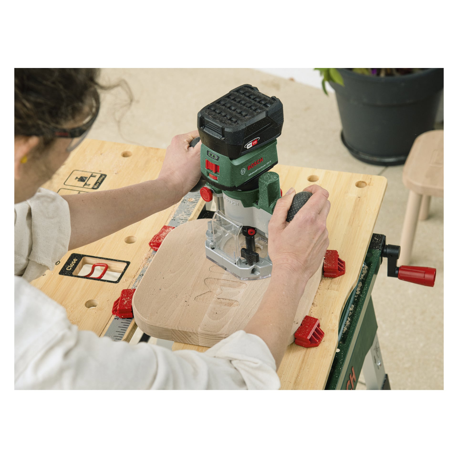 Jemand bedient den grün-schwarzen Bosch Power 4 All Combination Router AdvancedTrimTrimRouter 18V-8 mit Fräs-Tauchbasis, indem er eine wellenförmige Rille in eine leichte Holzplatte schneidet, die mit roten Klemmen an einer Werkbank befestigt ist. Im Hintergrund steht eine Topfpflanze und ein Holzhocker.