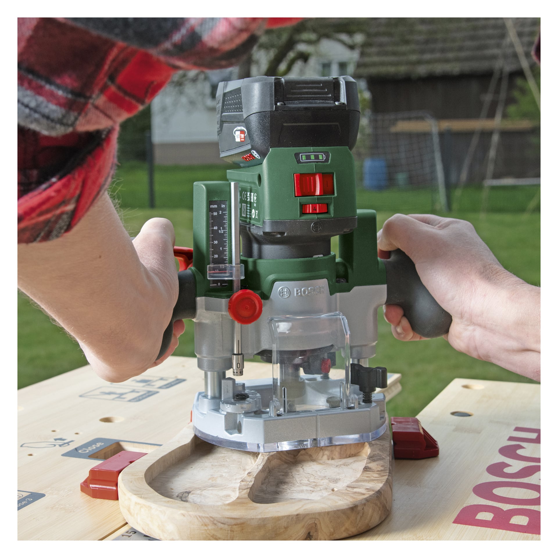 Jemand bedient den Bosch Power 4 All Combination Router AdvancedTrimRouter 18V-8 mit einer Fräser-Tauchbasis, wobei eine Holzplatte auf einer Außenwerkbank geformt wird. Die Person hält beide Griffe des Werkzeugs fest, während das Tablett mit roten Klammern fixiert wird, und im Hintergrund ist ein Haus mit Garten zu sehen.
