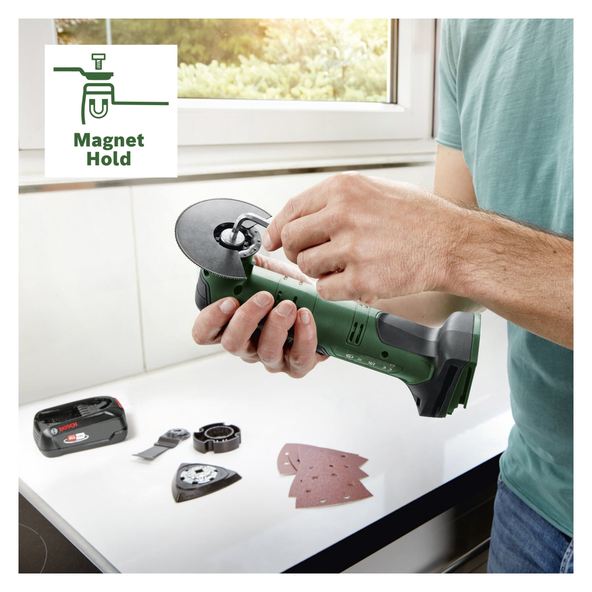 En person monterar en metallskiva i det gröna Bosch Power 4 All UniversalMulti 18V-32 verktygssetet på en bänkskiva, omgiven av verktygstillbehör, sandpappersbitar och ett batteri. En "Magnet Hold"-ikon syns uppe till vänster.