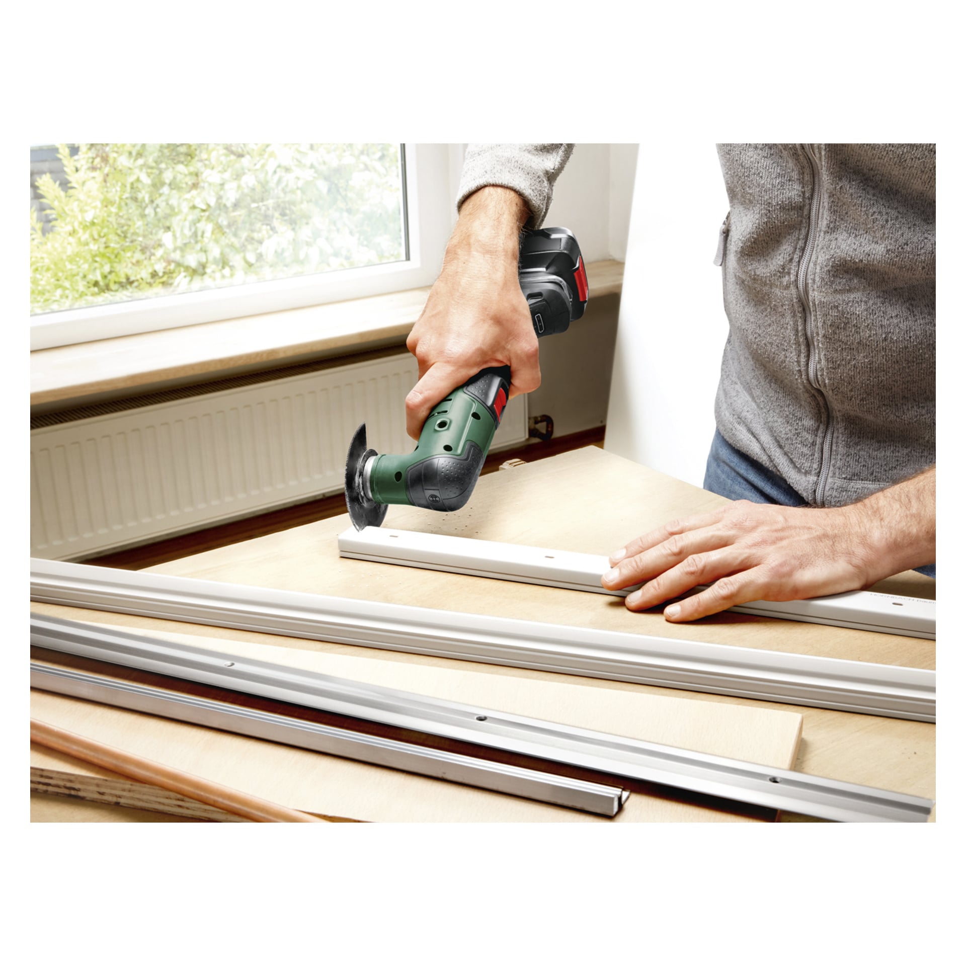 Noen bruker et grønt Bosch Power 4 All UniversalMulti 18V-32 multifunksjonsverktøy for å kutte en hvit stripe, muligens metall eller plast, på en arbeidsbenk nær et vindu, med ekstra lister og treplater spredt rundt.