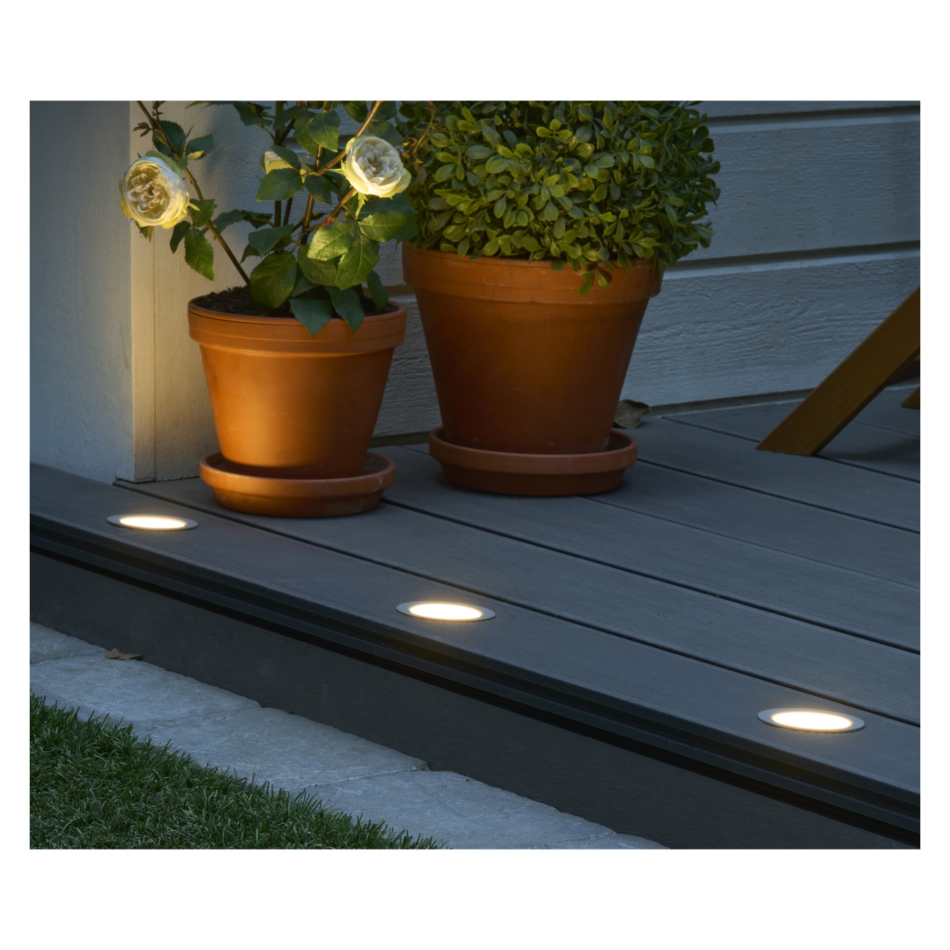 Zwei Topfpflanzen stehen auf einer grauen Terrasse neben der Hauswand. Drei runde Anslut Decklights Connectable System (4-Pack)-Lichter sind im Deckboden eingebettet und werfen Licht entlang der Kante. Die linke Pflanze zeigt gelbe, blumenförmige Lichter als Dekoration. Gras säumt den Rand des Decks.