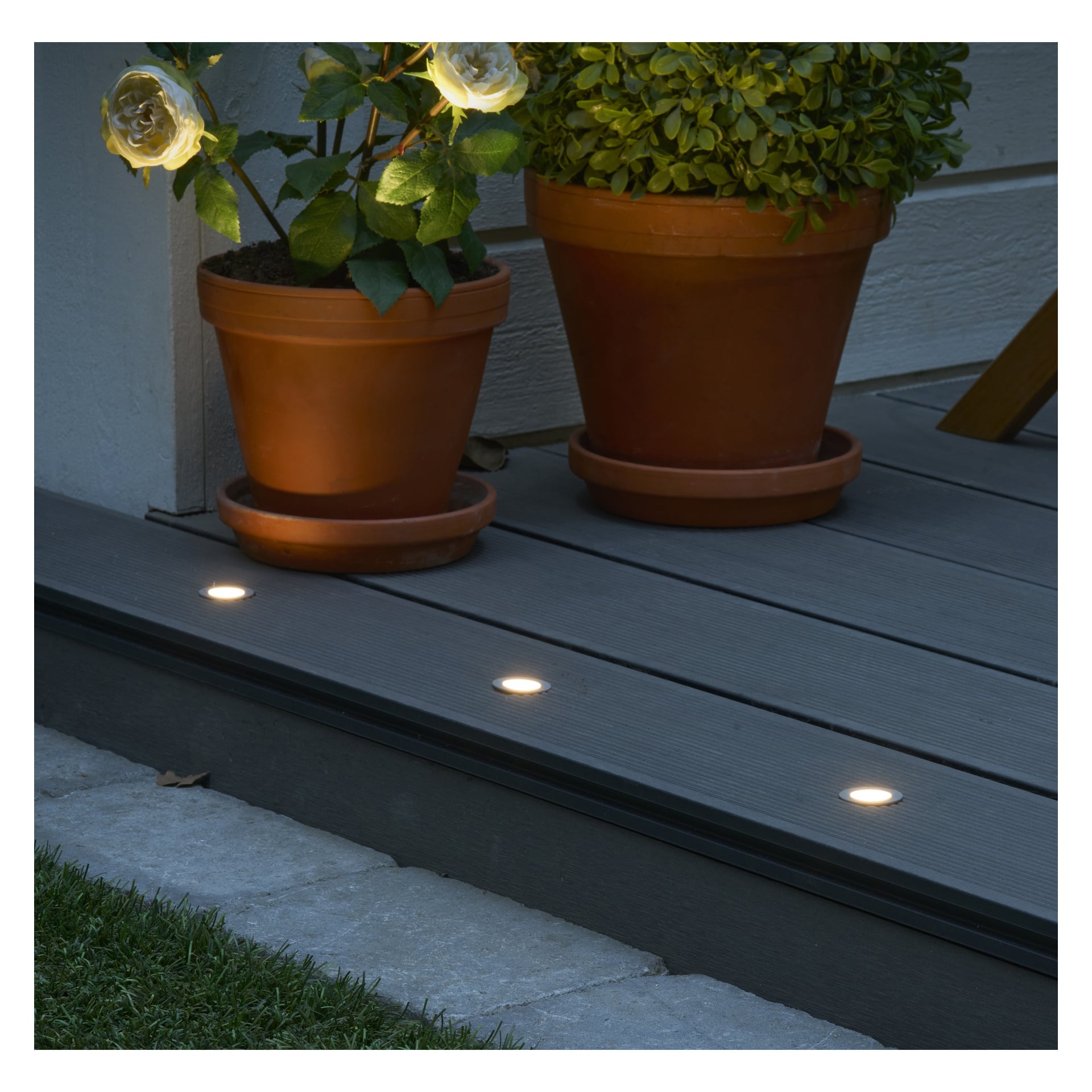 Fire runde Anslut Decklights Connectable System LED-lys er plassert i en grå treterrasse ved siden av to potteplanter. Deres myke glød lyser forsiktig opp området rundt om kvelden eller natten.