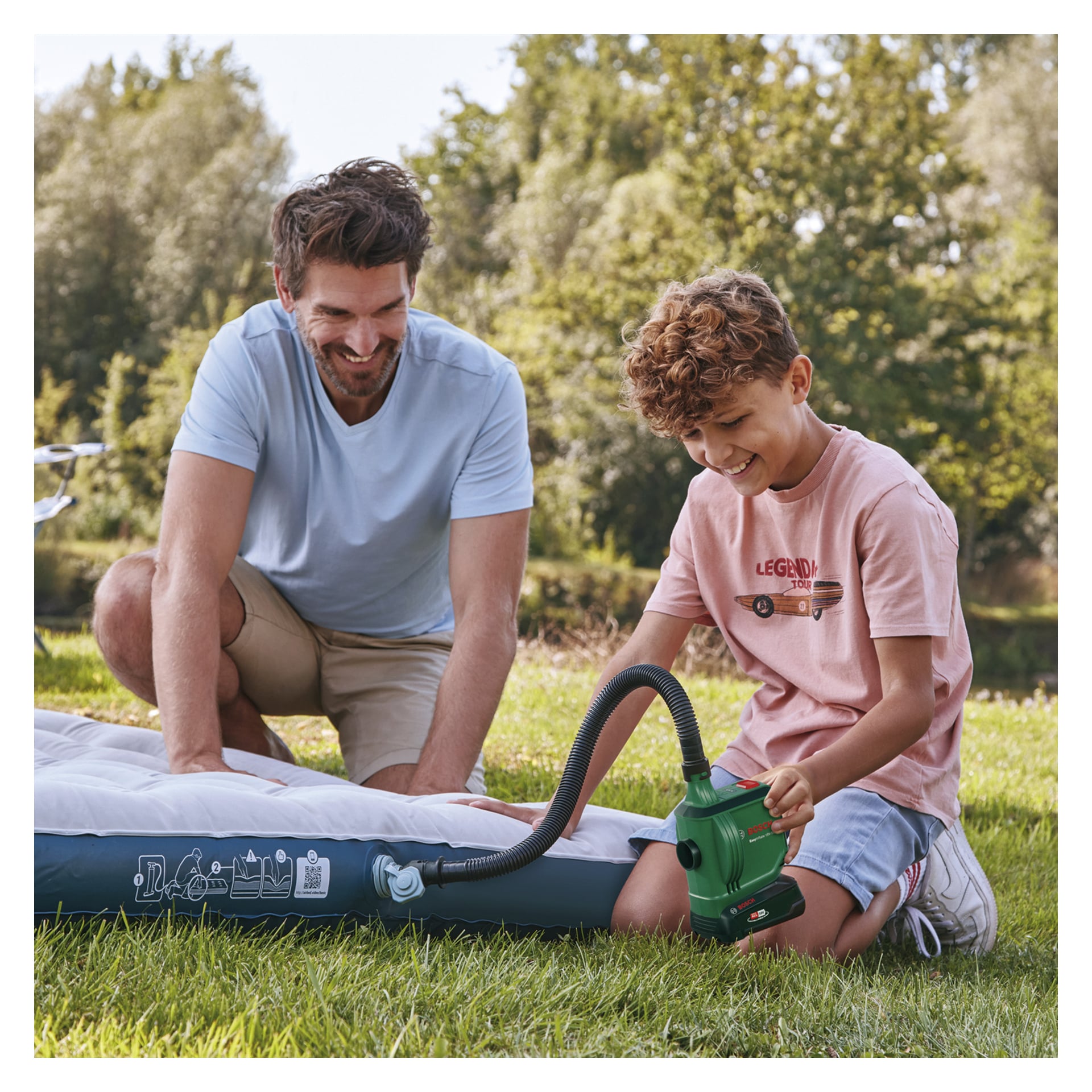 Ein Mann und ein Junge sitzen draußen auf dem Gras und pumpen eine Luftmatratze mit einer grünen Bosch Power 4 All EasyInflate 18V-500 batteriebetriebenen Luftpumpe auf. Der lächelnde Junge hält die Zapfsäule, während der Mann neben ihm kniet und ebenfalls lächelt. Im Hintergrund stehen Bäume.