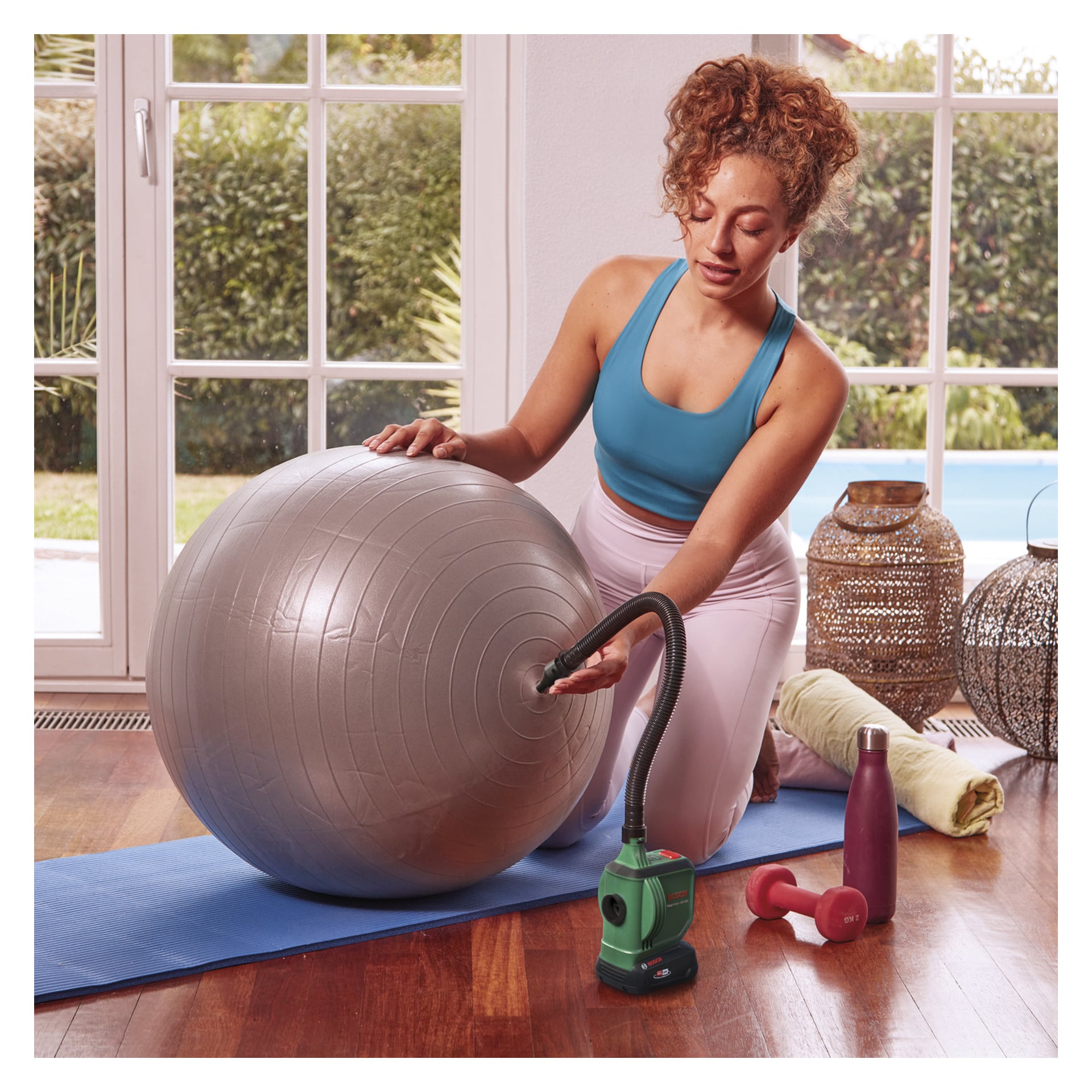 Eine Frau in Sportkleidung kniet auf einer Yogamatte im Inneren und bläst einen grauen Trainingsball mit der Bosch Power 4 All EasyInflate 18V-500 akkulosen Luftpumpe auf. Neben ihr liegen eine Wasserflasche, zwei Hanteln und ein zusammengerolltes Handtuch. Große Fenster hinter ihr zeigen grüne Bäume draußen.