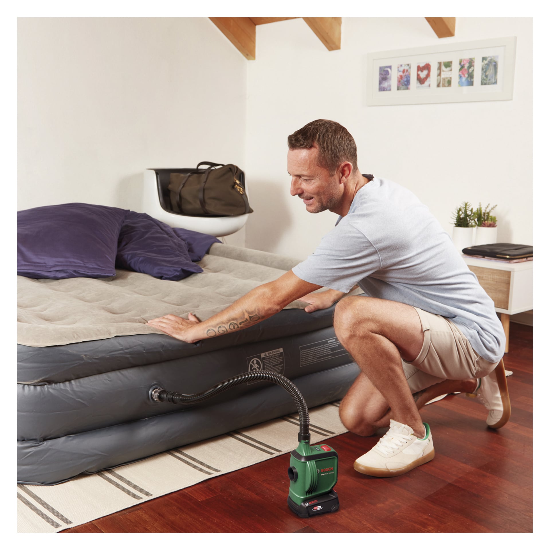 Ein Mann kniet auf dem Schlafzimmerboden und bläst eine graue Luftmatratze mit der Bosch Power 4 All EasyInflate 18V-500 kabellosen Luftpumpe auf. Das Bett im Zimmer hat zwei lila Kissen, und auf einem nahegelegenen Stuhl liegt eine Tasche.