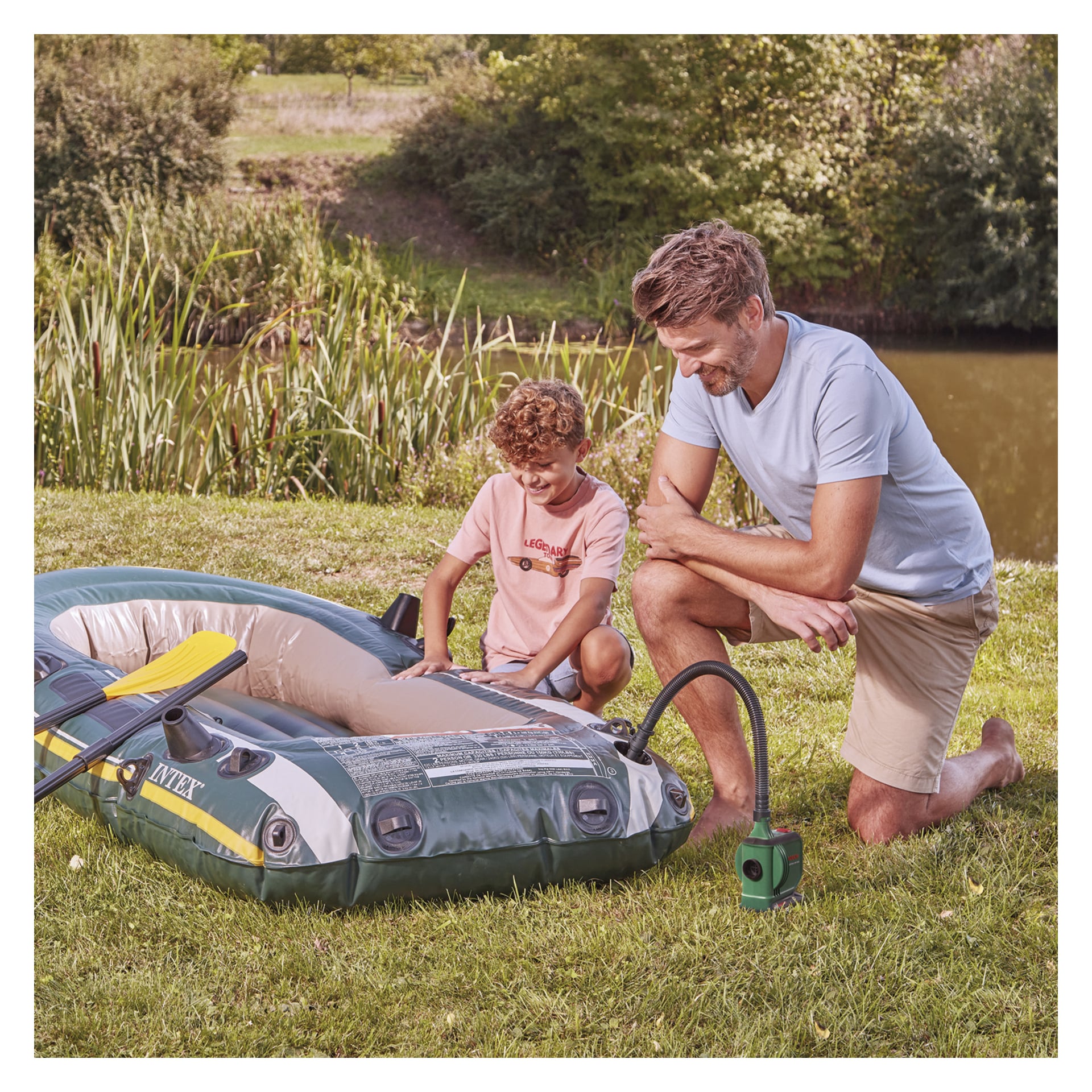 Ein Mann kniet auf dem Gras neben einem aufblasbaren Boot und benutzt eine Bosch Power 4 All EasyInflate 18V-500 Luftpumpe. Ein Junge steht in der Nähe und beobachtet ihn, mit einem Teich und Bäumen im Hintergrund.
