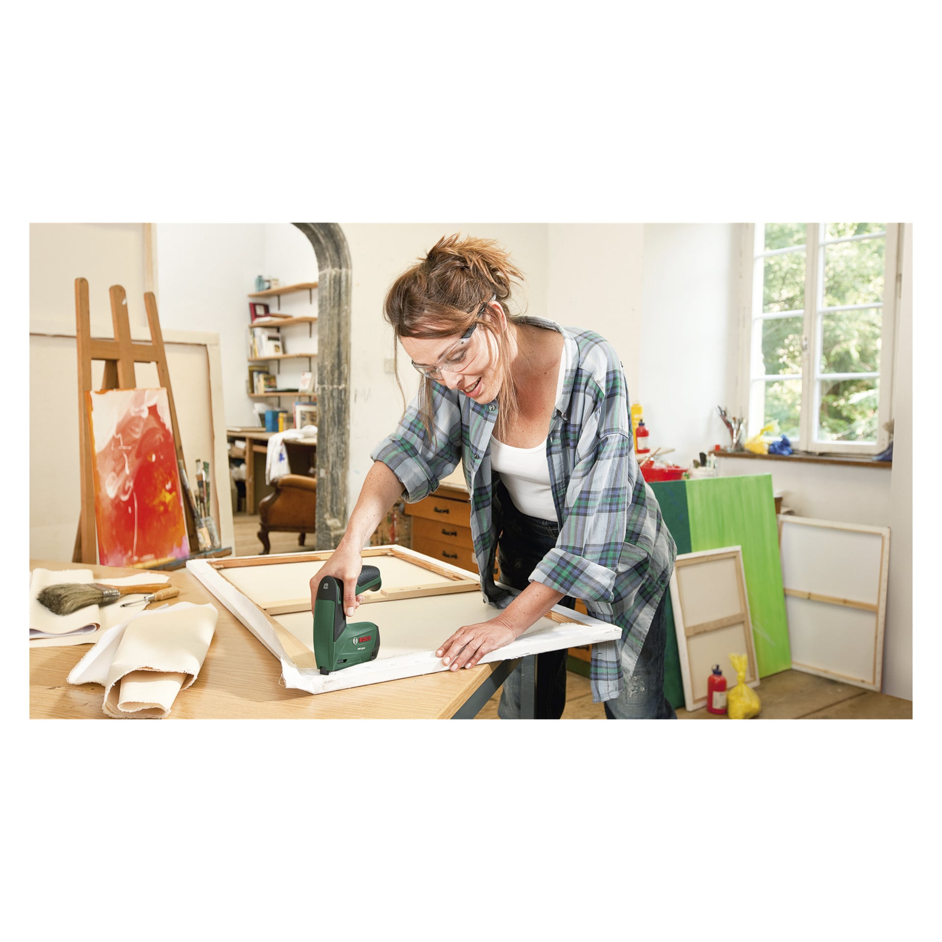 Eine Frau mit Brille befestigt in einem Kunststudio eine Leinwand an einem Holzrahmen mit der Bosch PTK 3,6 LI batteriebetriebenen Tackerpistole. Der Hintergrund zeigt mehrere Leinwände, verschiedene Kunstmaterialien und ein Fenster, das das Grün draußen zeigt.